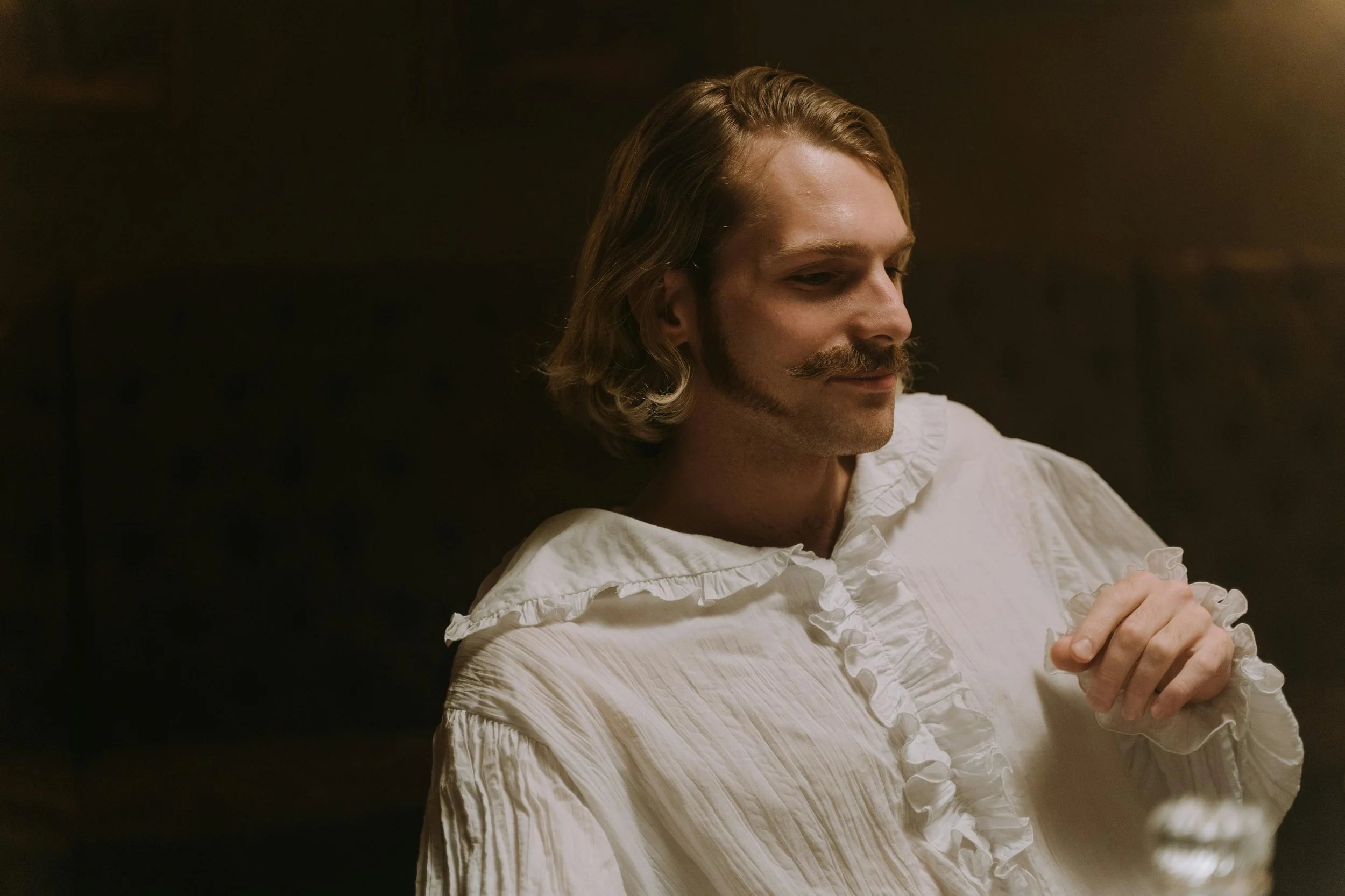 A man with shoulder-length blond hair, a mustache, and a beard is wearing a white ruffled shirt and sitting in a dimly lit room.