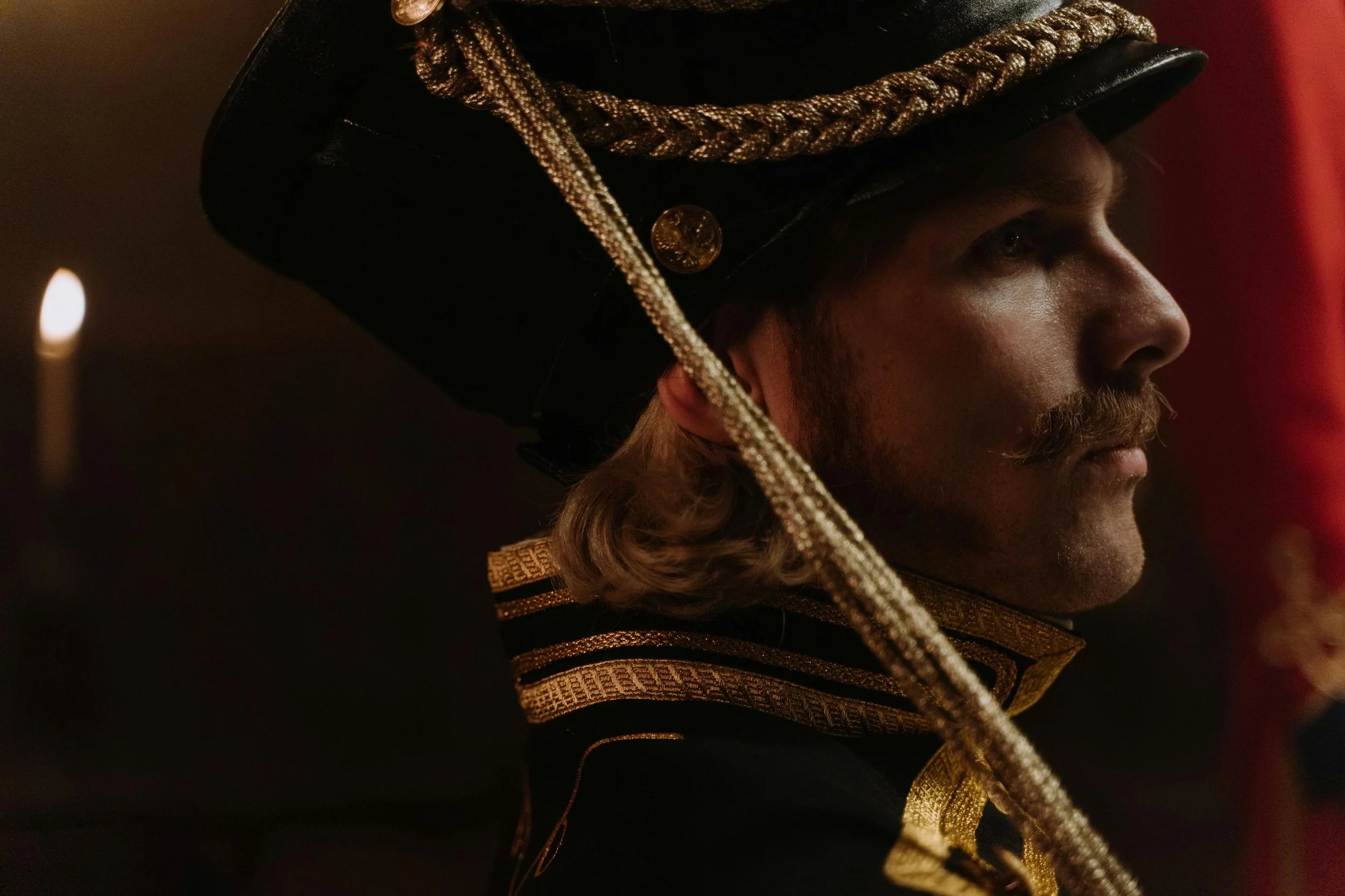 Close-up side profile of a man in a historical military uniform, wearing a tall black hat with gold braid, epaulet, and a gold button, in a dimly lit setting.
