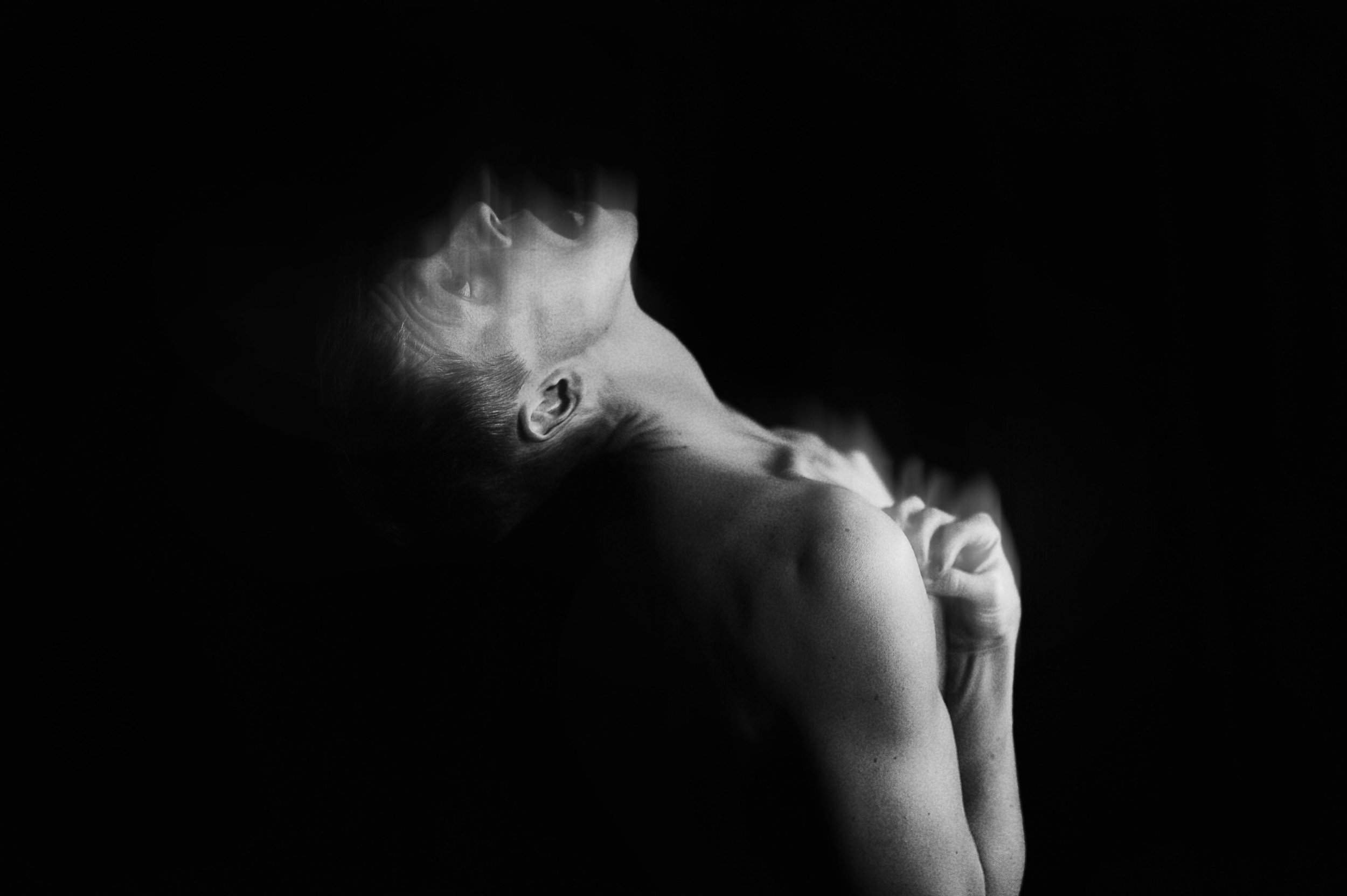 A black and white photograph of a man with his head tilted back, eyes closed, shrieking, and hands clasped near his chest, with dramatic lighting emphasizing his shoulder and face against a dark background.