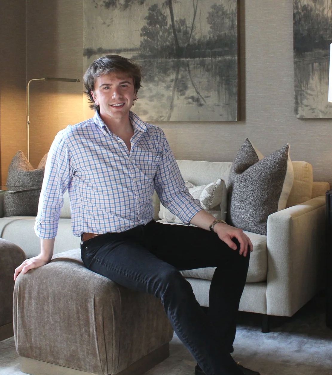A young man with brown hair, wearing a checkered shirt and black pants, is sitting on an ottoman in a living room, smiling at the camera. There is a beige sofa with decorative pillows and wall art with a nature scene behind him.