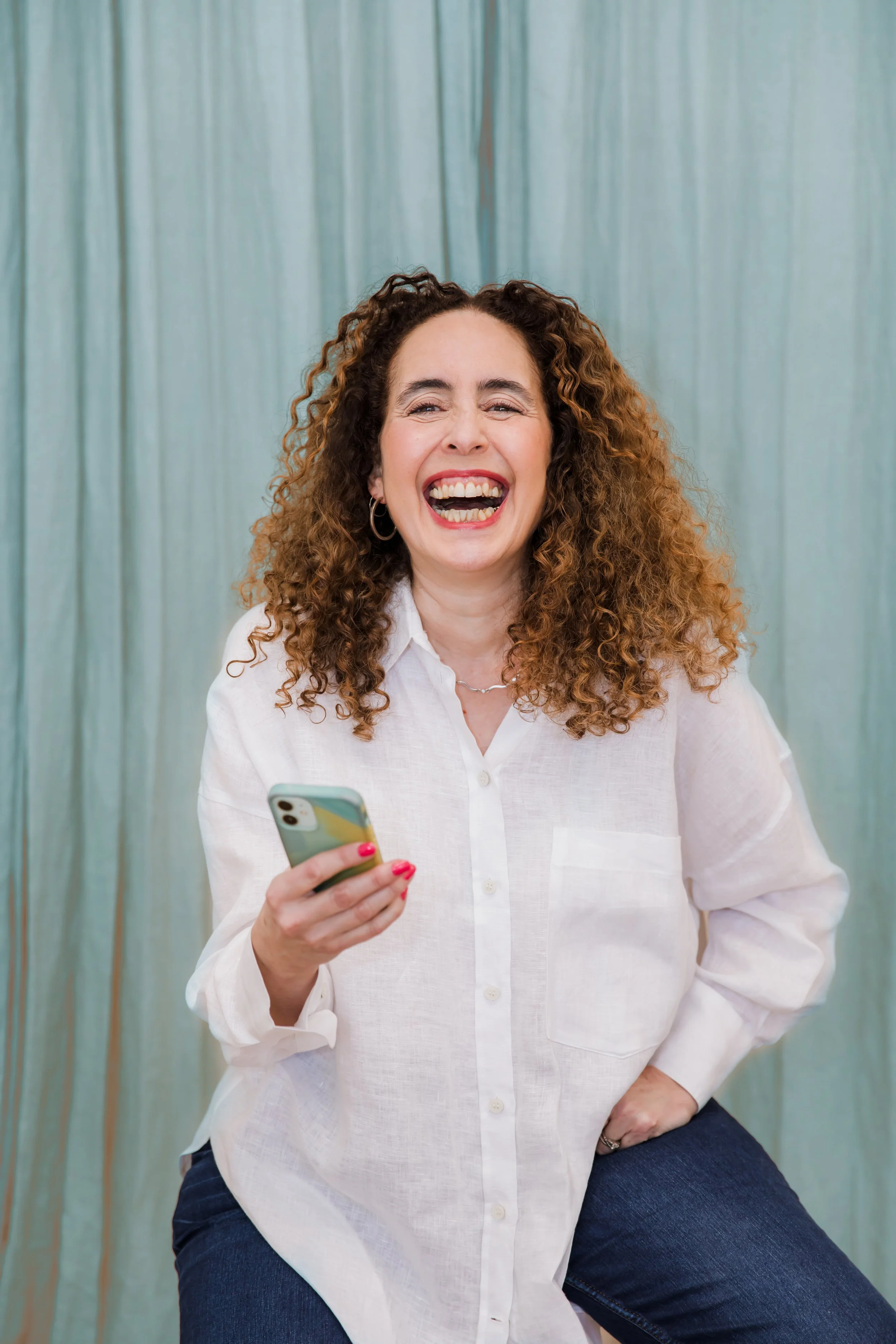 Woman with curly brown hair laughing while holding a smartphone, wearing a white shirt and jeans, in front of a turquoise curtain background.