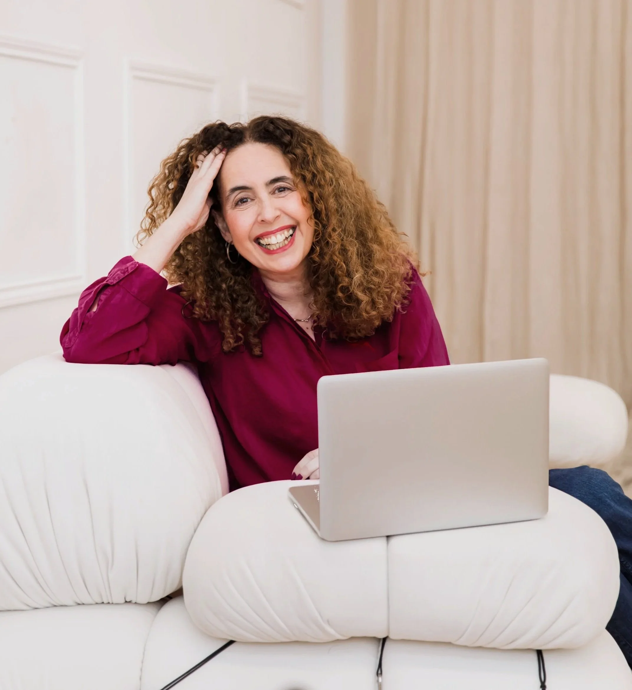 Woman with curly hair smiling, sitting on a white couch, using a silver laptop.