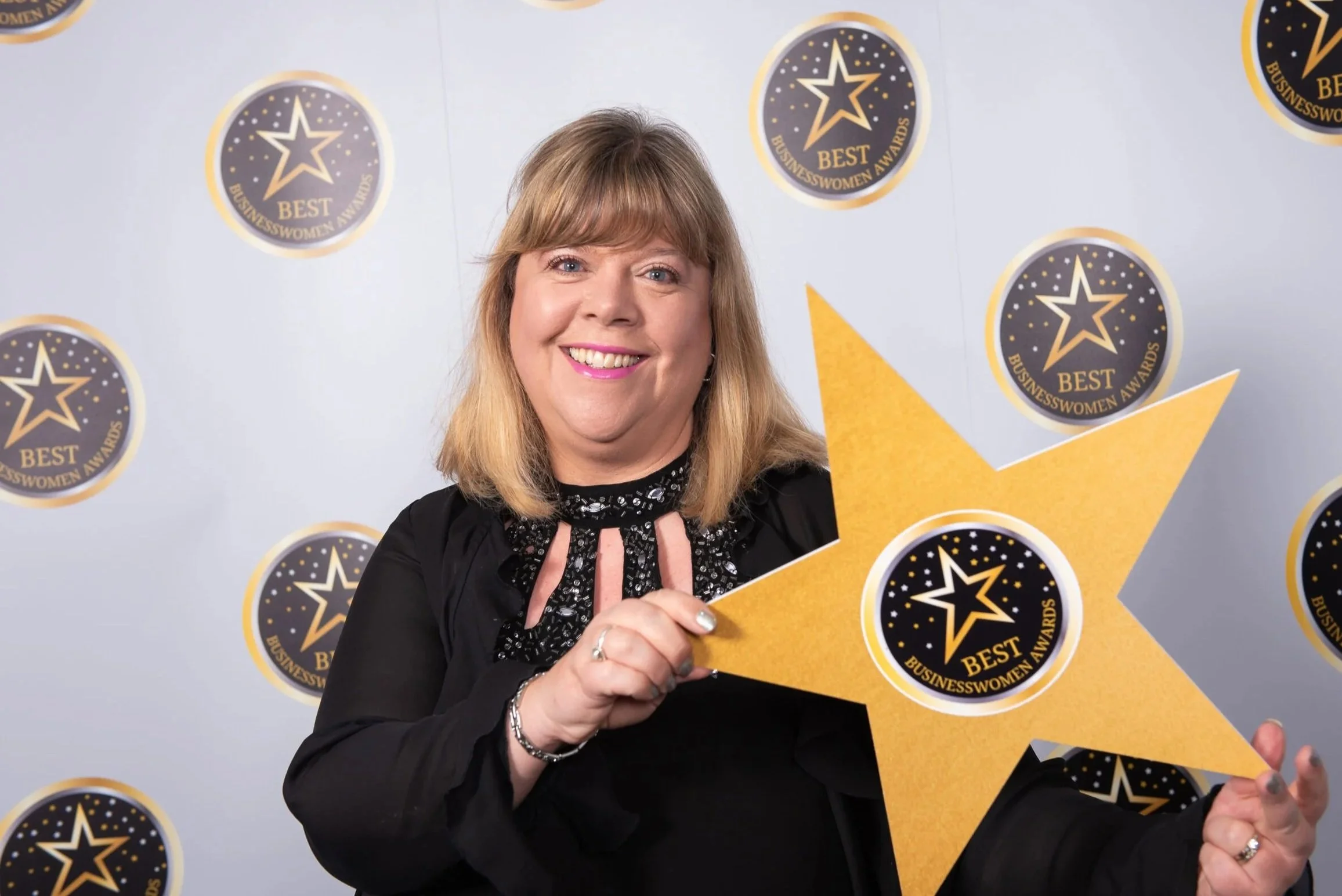 A woman with blonde hair smiling at the camera, holding a large yellow star with a "BEST BUSINESSWOMEN AWARDS" logo on it, standing in front of a backdrop with multiple similar logos.