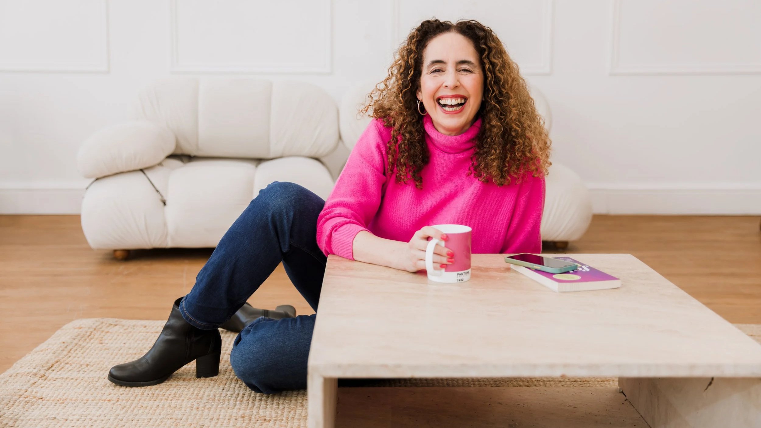A woman with curly hair, wearing a bright pink sweater, jeans, and black ankle boots, sitting on the floor and leaning on a low wooden coffee table. She is smiling and holding a white mug, with a smartphone and a book on the table in front of her. There is a white sofa behind her in a room with wooden flooring.