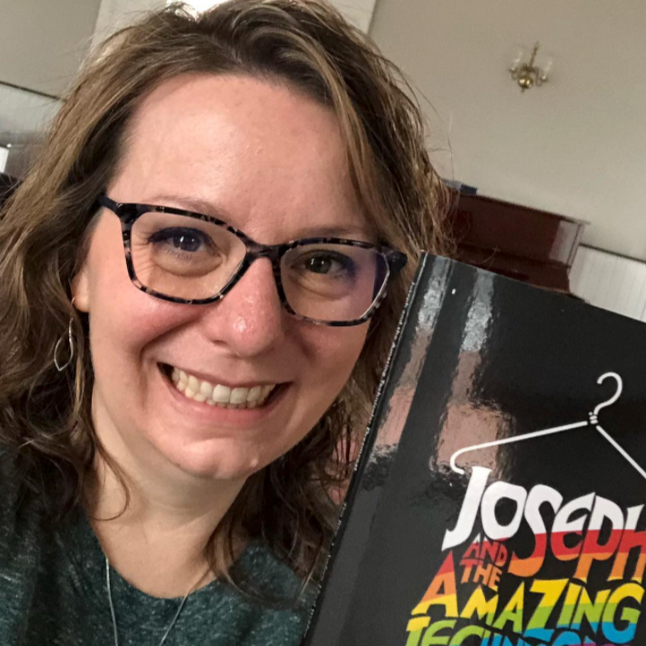 Woman smiling and holding a book titled 'Joseph and the Amazing Technicolor Dreamcoat' indoors.