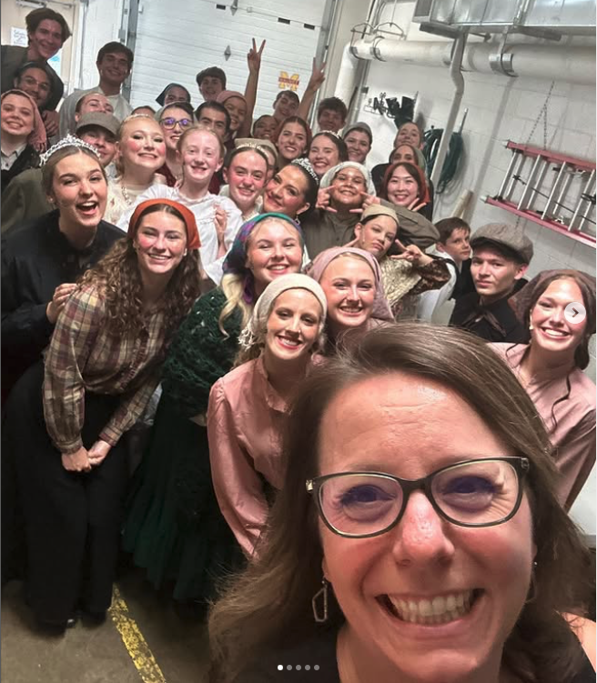 Group of smiling young people and adults gathered indoors, with some making peace signs, in a large room with industrial pipes and equipment in the background.