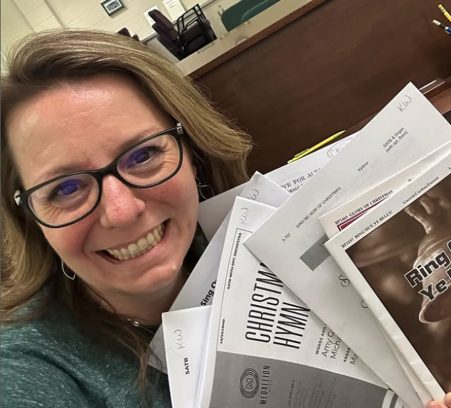 A woman smiling wearing glasses, holding several sheets of paper or scripts, in an office setting.