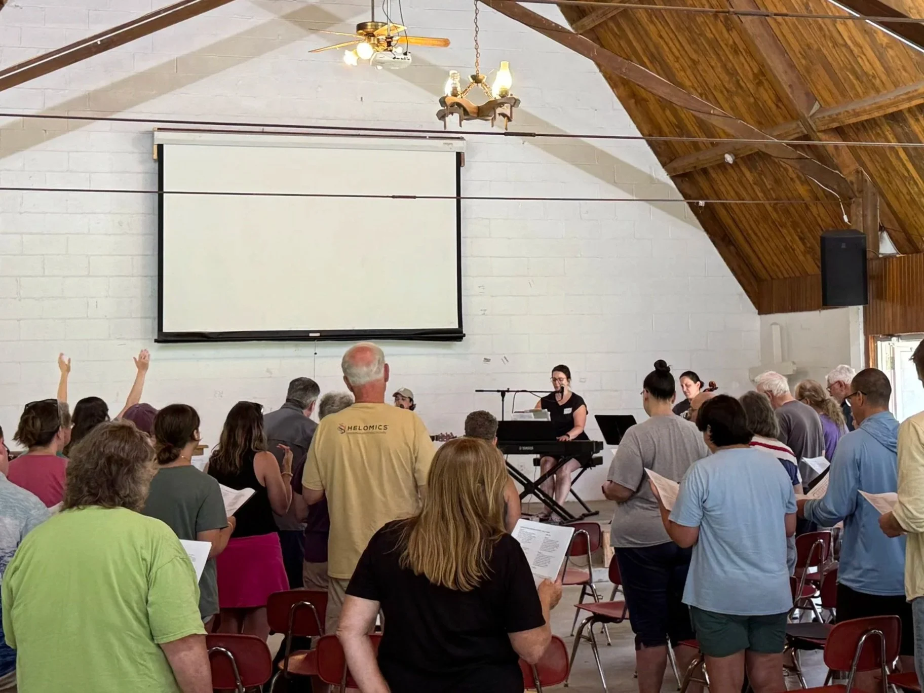 People attending a church service in a wooden church with vaulted ceiling, ceiling fans, and a large white wall with a projection screen at the front.