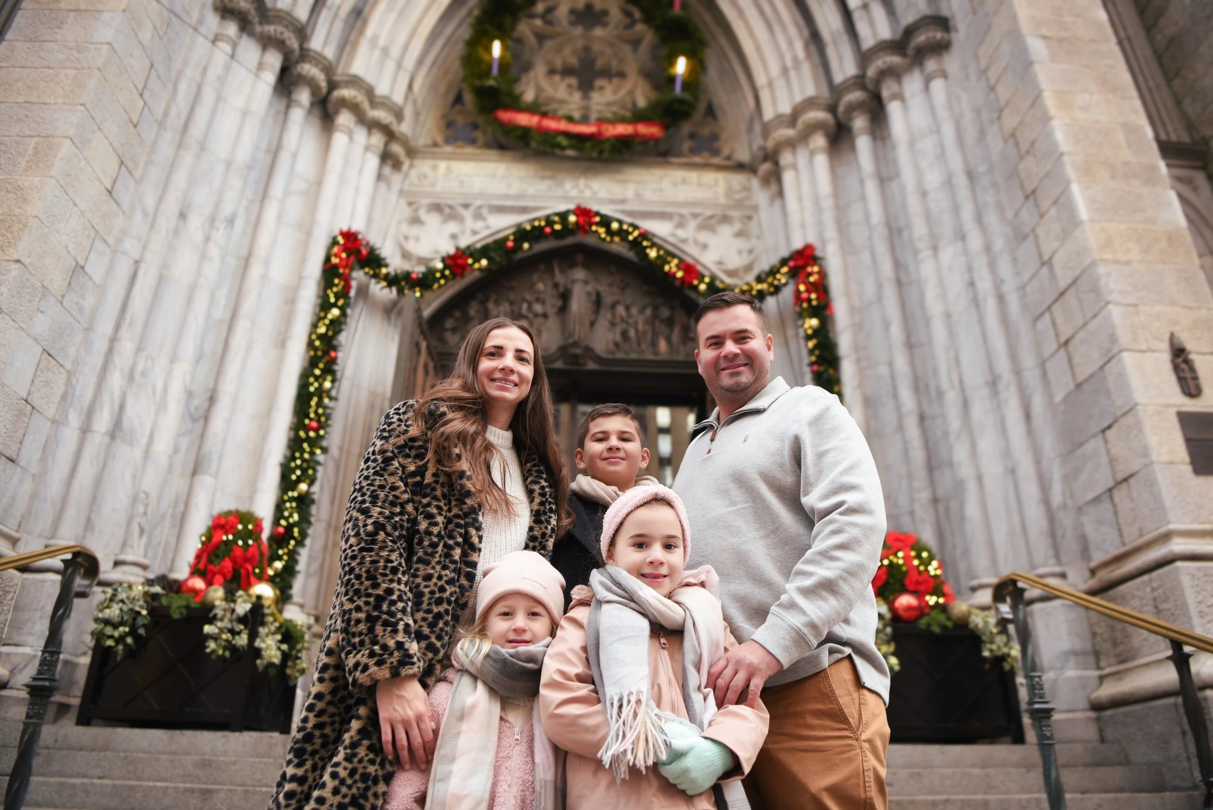 A family of five smiling and posing in front of St. Patrick's Cathedral  decorated with Christmas wreaths and garlands.