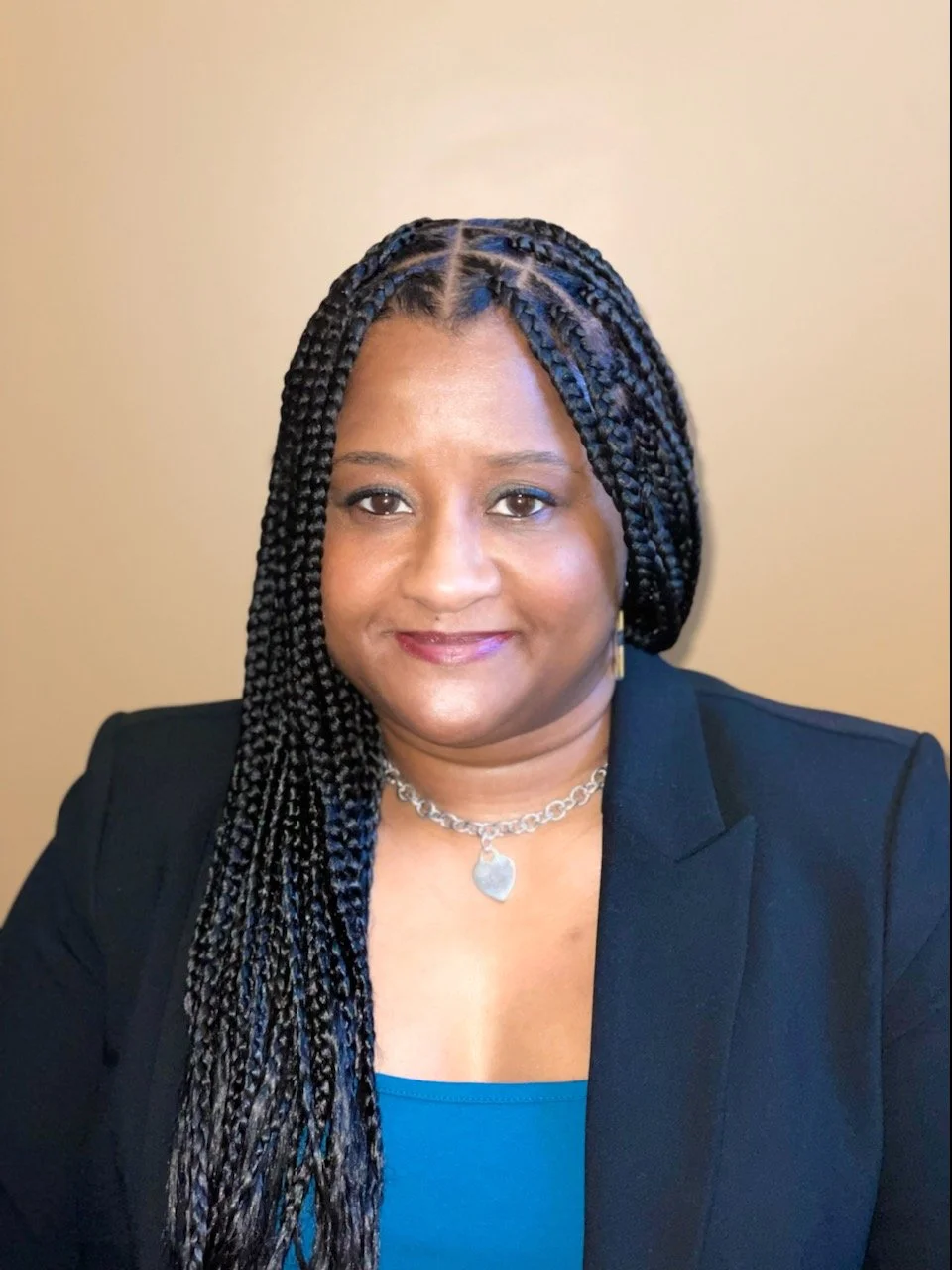 A woman with braided hair wearing a black blazer, a blue top, and a silver heart necklace, smiling against a light beige background.