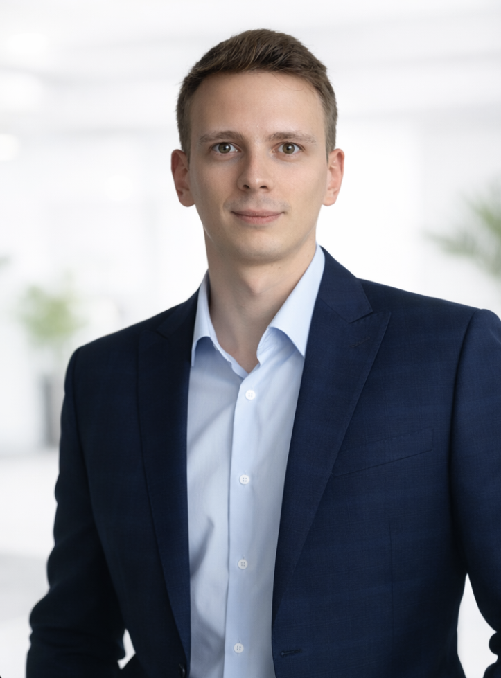 A young man in a navy blue suit and white dress shirt standing in a bright, modern office environment.