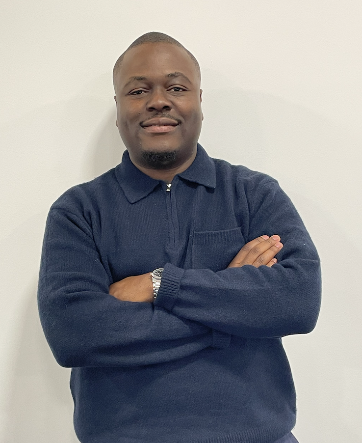 A man with a dark skin tone and short hair standing against a plain white wall, wearing a navy blue sweater with a zipper and a watch on his left wrist, smiling with arms crossed.