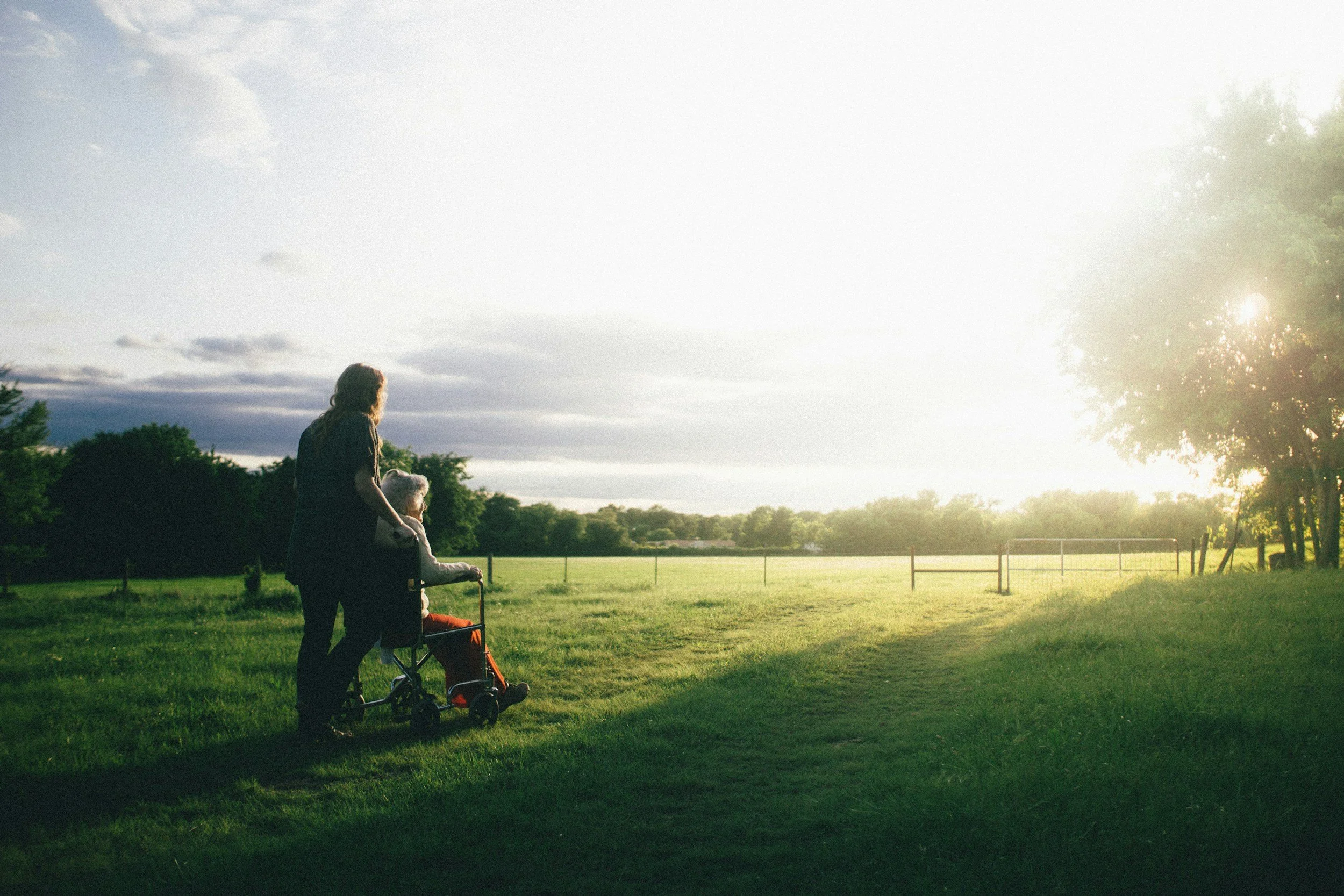 a lady in a wheelchair looking out onto a field at sunset with her carer standing behind her