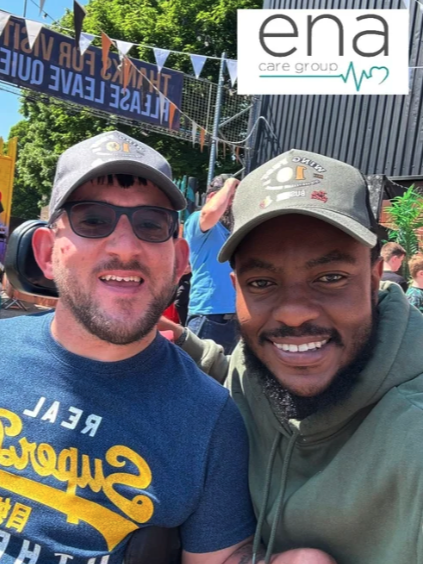 Two smiling men posing for a selfie at an outdoor event, with ENA Care Group branding in the top corner. Colourful bunting and event banners are visible in the background, creating a friendly, community atmosphere.