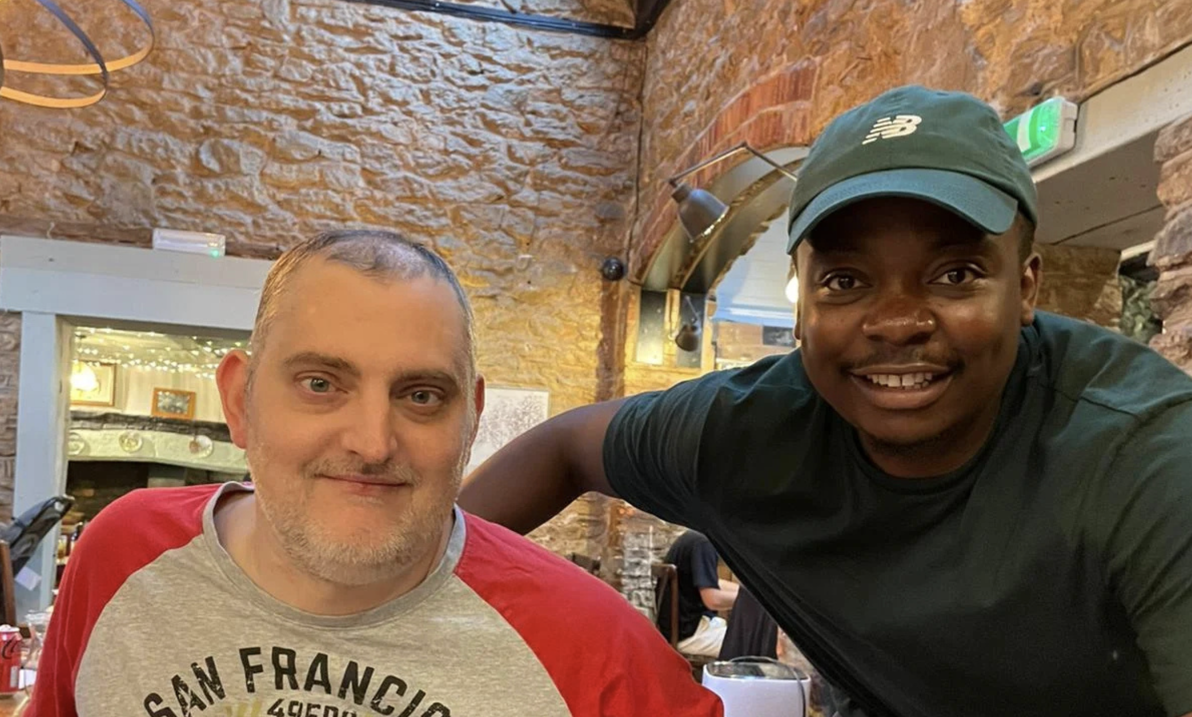 Two men sitting together in a restaurant with brick walls. The man on the left has short gray hair, facial hair, and is wearing a red and gray shirt. The man on the right, wearing a green cap and black shirt, is leaning over and smiling at the camera.