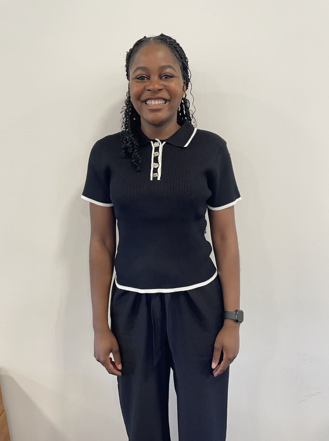 A young woman with dark curly hair and a bright smile, standing against a plain white wall, wearing a black short-sleeved polo shirt with white trim and black pants.