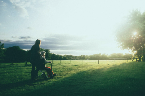 an elderly lady in a wheelchair overlooking a field at sunset with her carer standing behind her