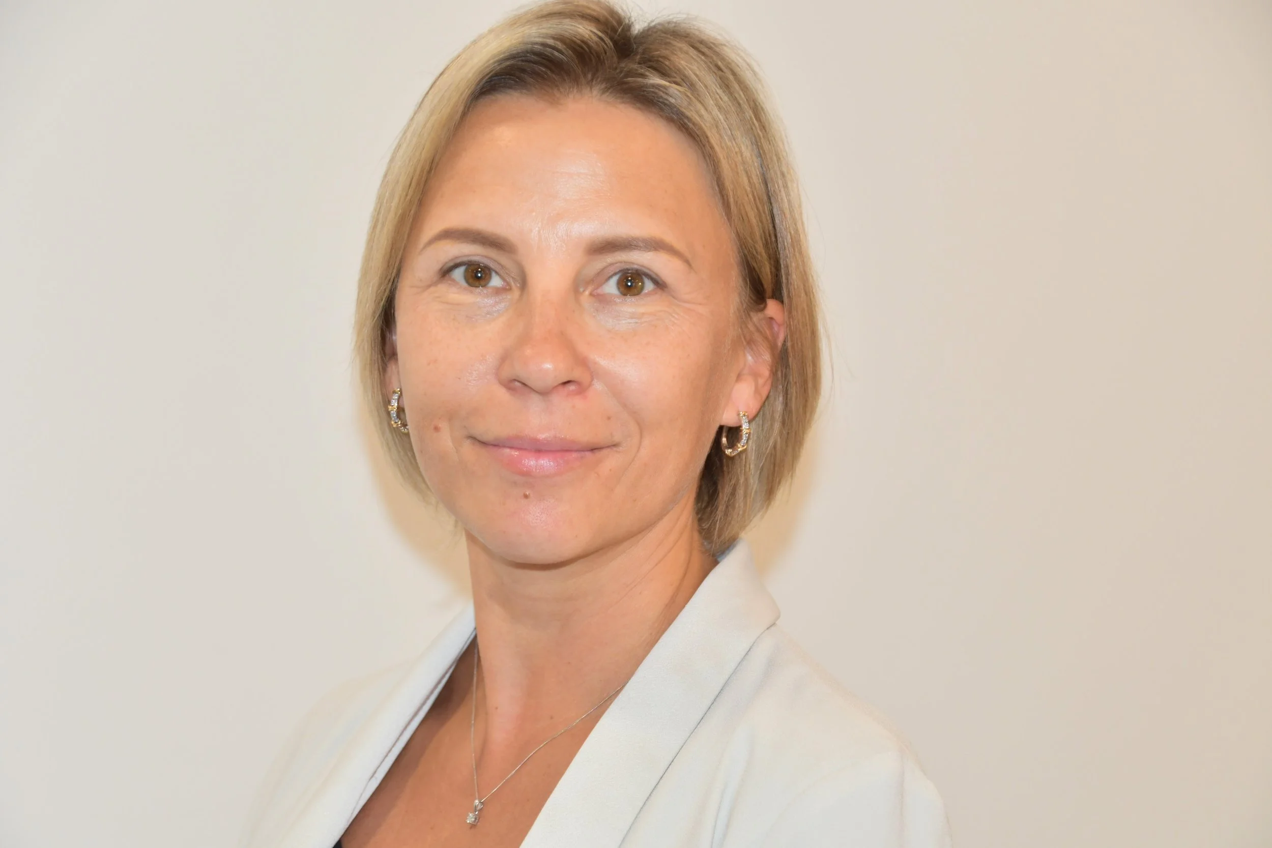 A middle-aged woman with short blonde hair, wearing a white blazer and jewelry, standing against a plain light-colored background.