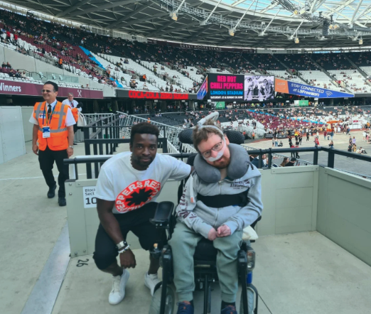 Two young men sitting together in a stadium, one in a wheelchair, with a large sports stadium background and digital screens showing event information.