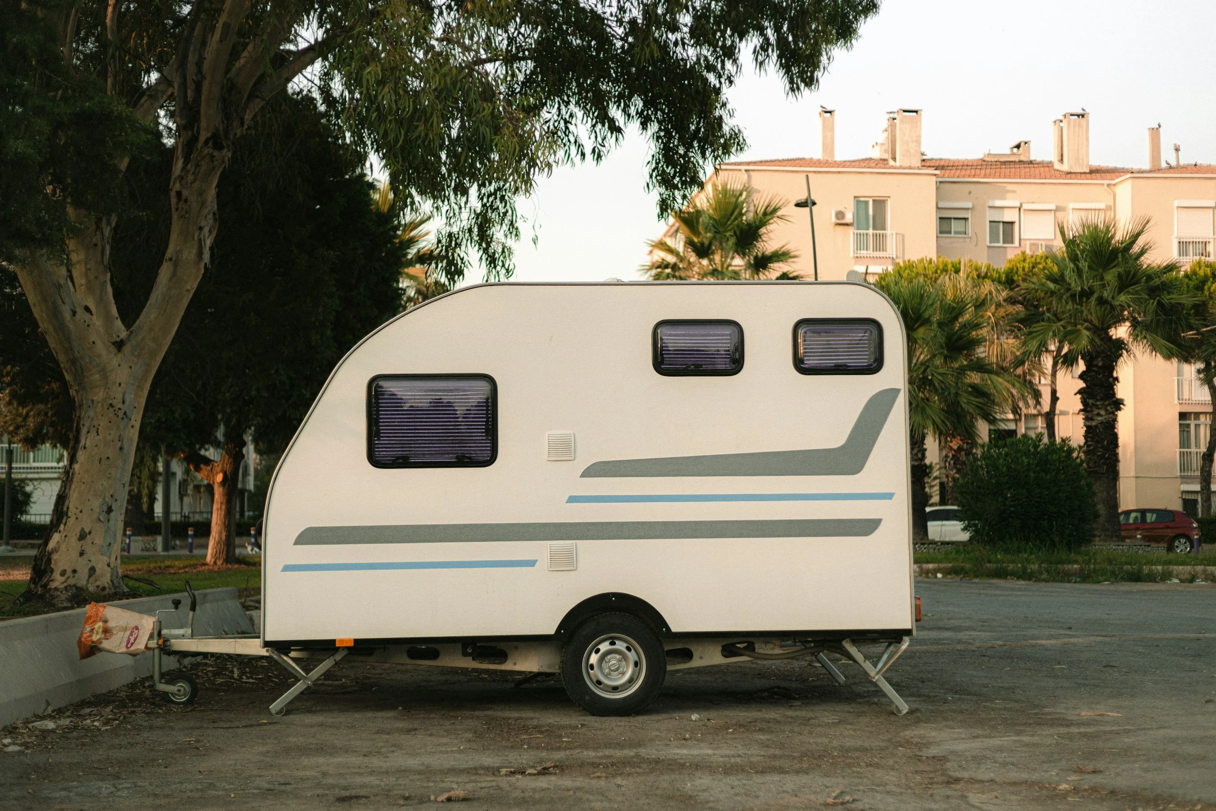 En hvit campingvogn parkert på en tom parkeringsplass med trær og boligblokker i bakgrunnen.