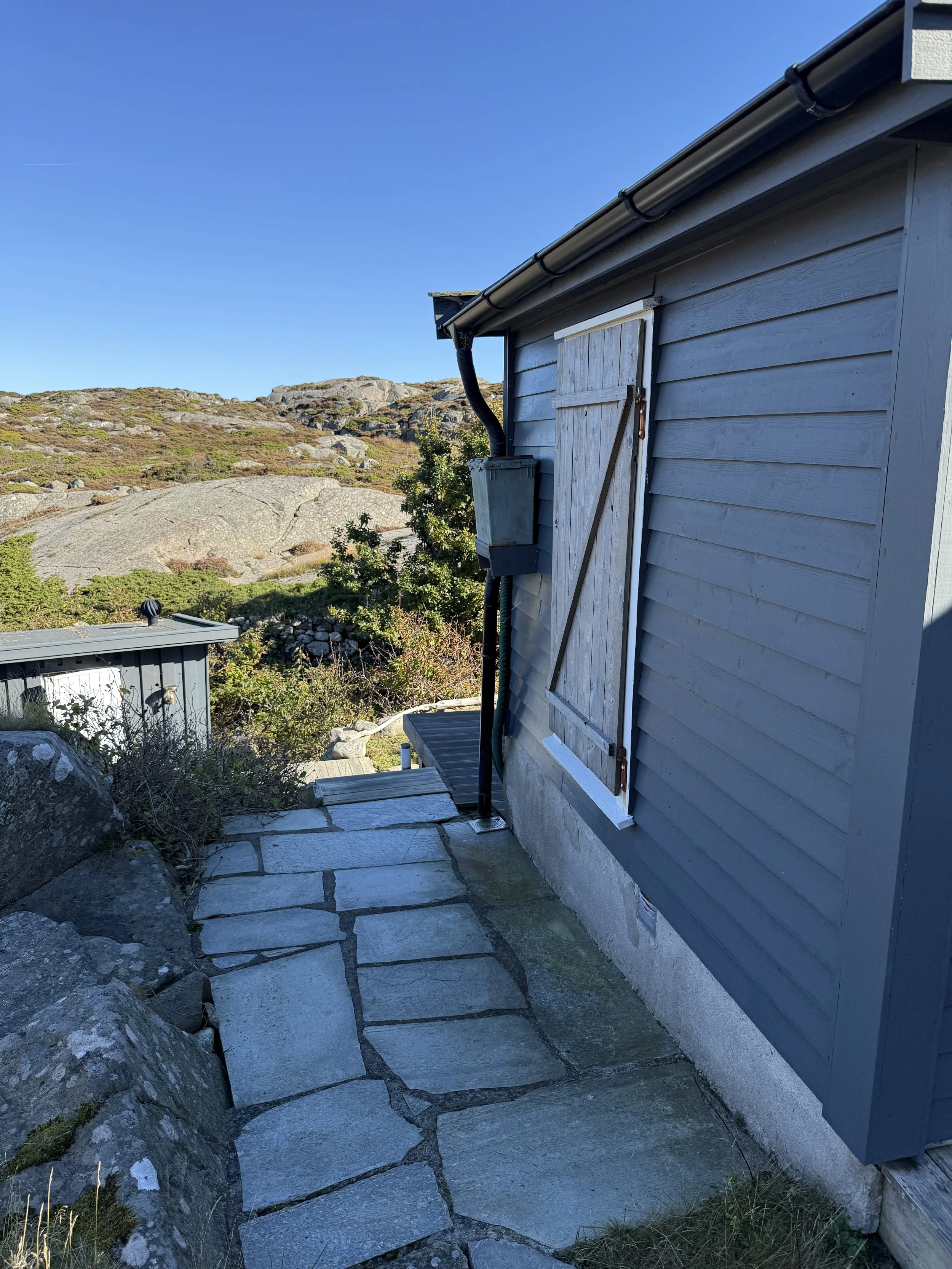 Utsikt over en blå husvegg med en avstengt vindusluke, omgitt av steintrapper og fjell i bakgrunnen under klar blå himmel.