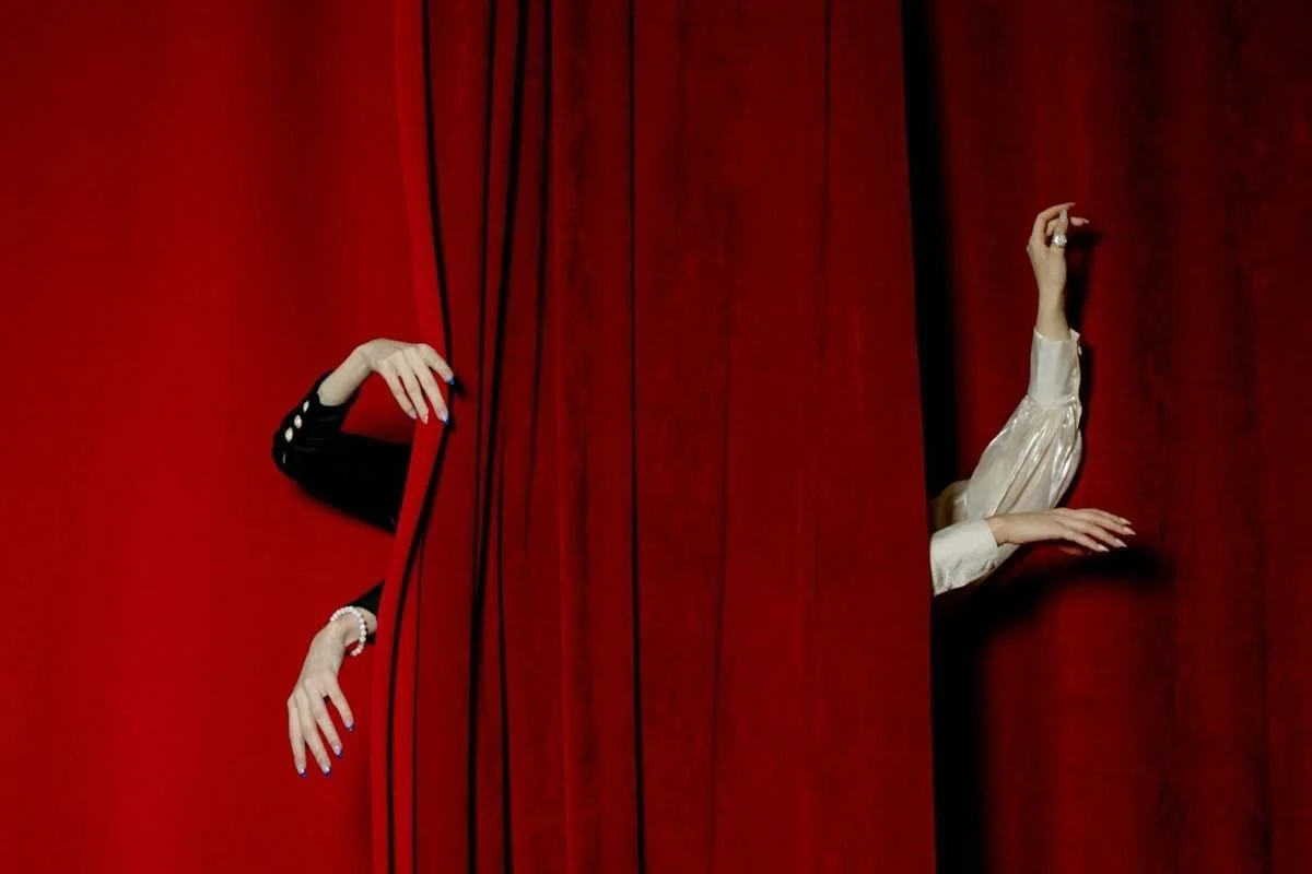 Two women are peeking out from behind red theater curtains, with only their hands and arms visible.