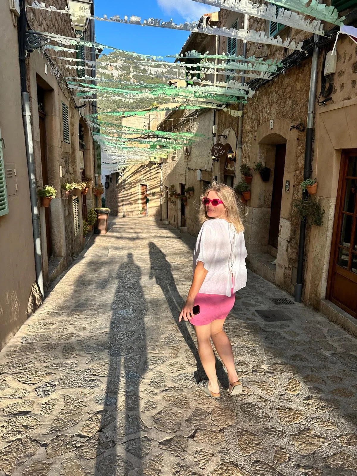 A woman with blonde hair wearing pink sunglasses, a white top, and a pink skirt standing on a cobblestone street. She is holding a phone in her right hand and looking back at the camera. The street has hanging decorations overhead and stone buildings with flower pots along the walls.
