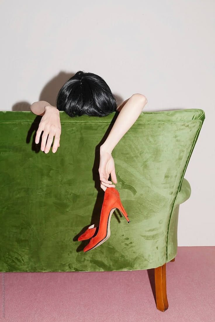 A person with black hair is sitting with their head and arms hanging over the back of a green velvet couch, holding a red high-heeled shoe.