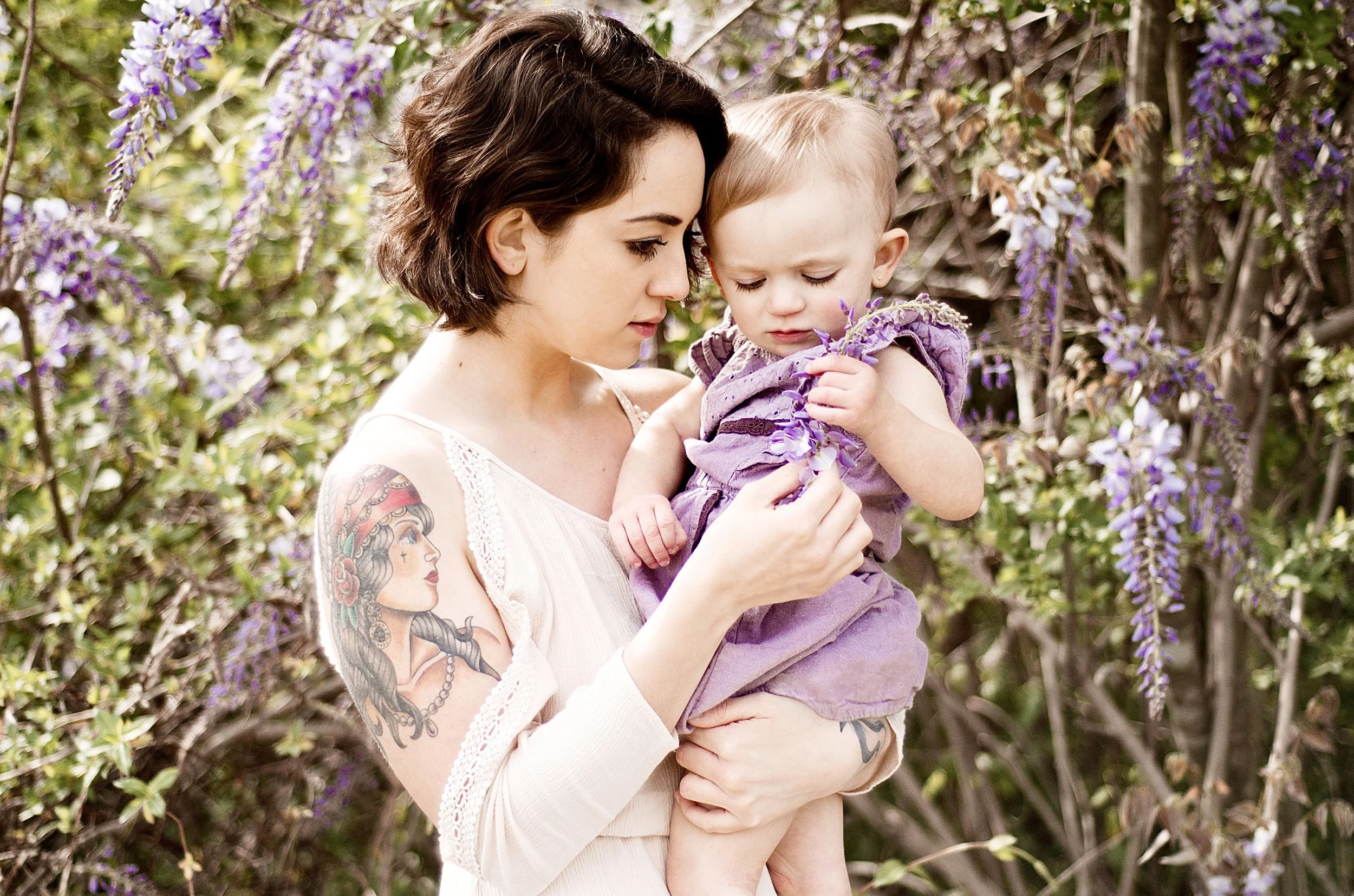 dark haired woman wearing white dress holds daughter in purple dress while looking at purple wisteria flower, clarksville tn photographer, nashville tn photographer, affordable, motherhood, lifestyle, editorial, bright, colorful, bold, neutral