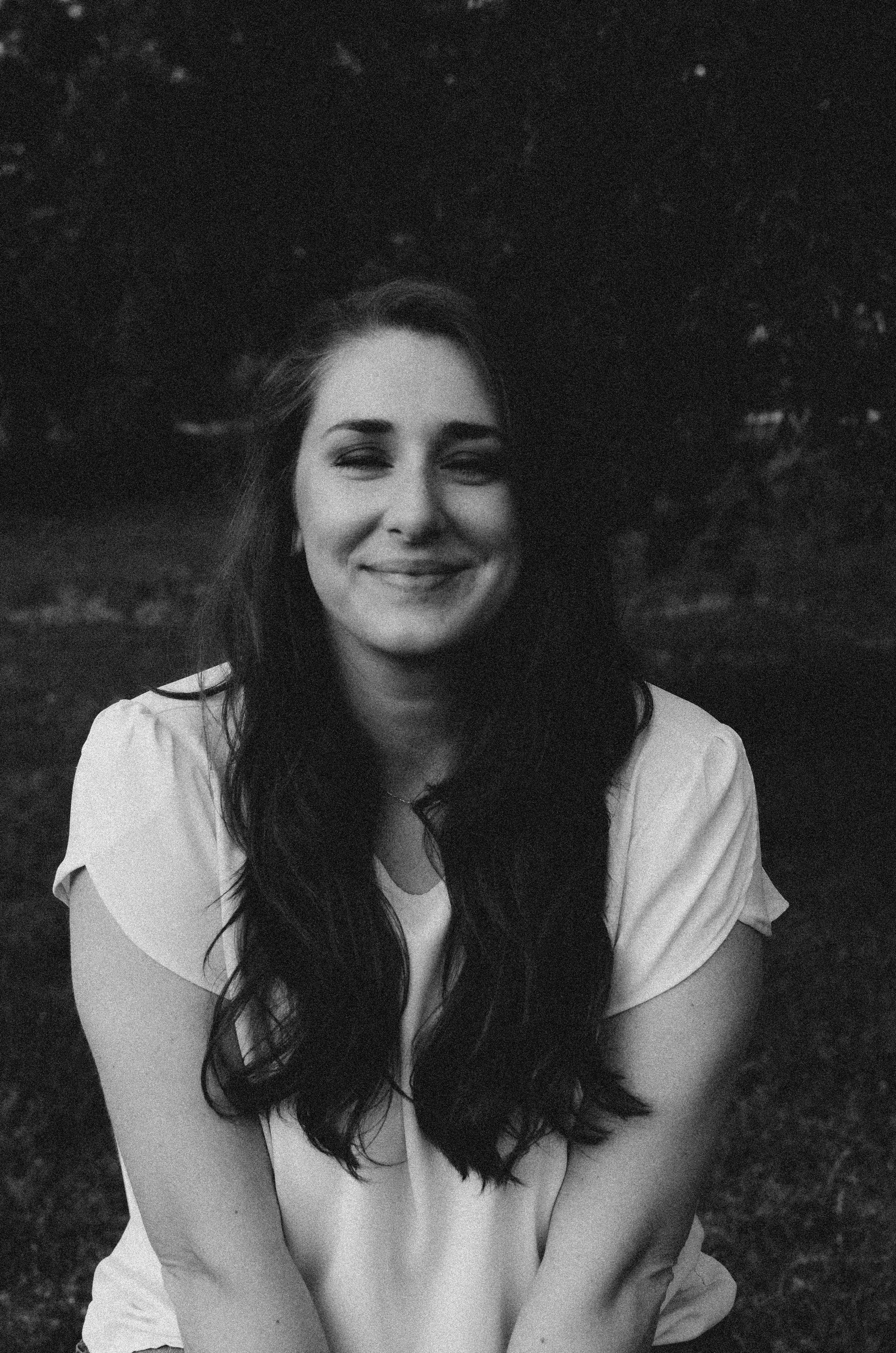 Black and white photo of a young woman with long dark hair, smiling with eyes closed, wearing a light-colored top, outdoors at sunset
