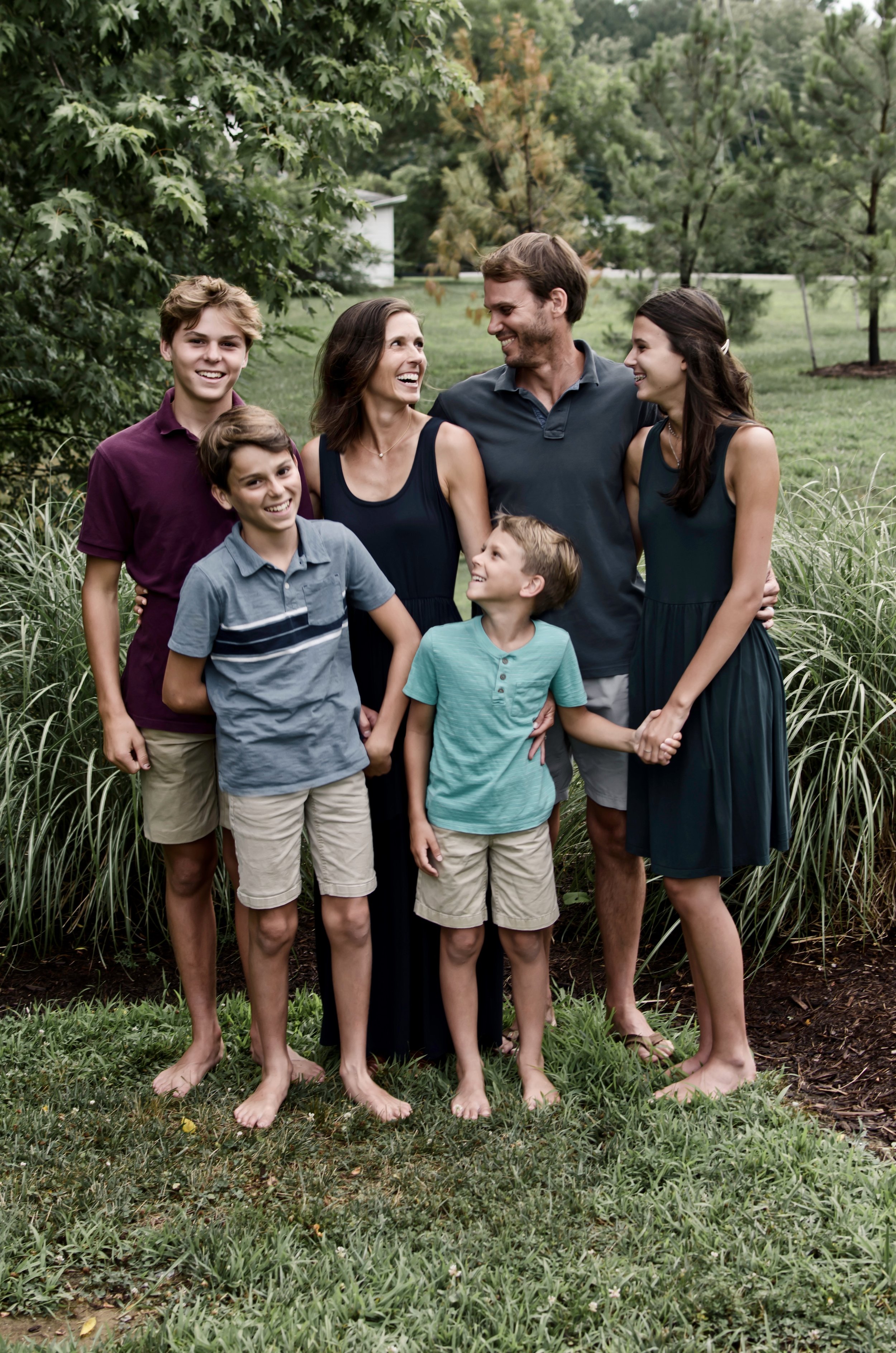 A family of seven standing outdoors barefoot on grass with trees and a lawn in the background. They are smiling and looking at each other, dressed in casual summer clothes.