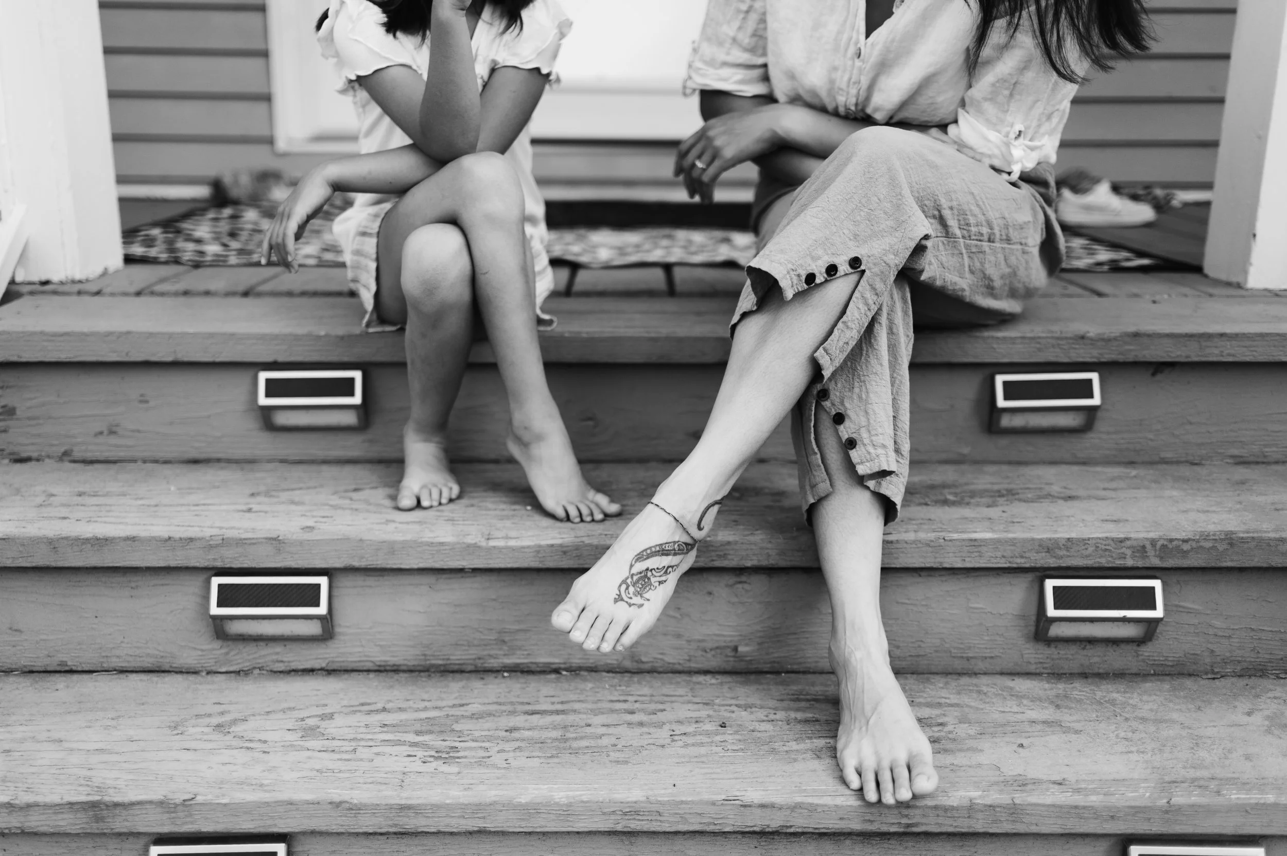 Black and white photo of a woman and a young girl sitting on wooden steps outside. They are sitting in an identical pose with their legs crossed and arms resting on knees. 