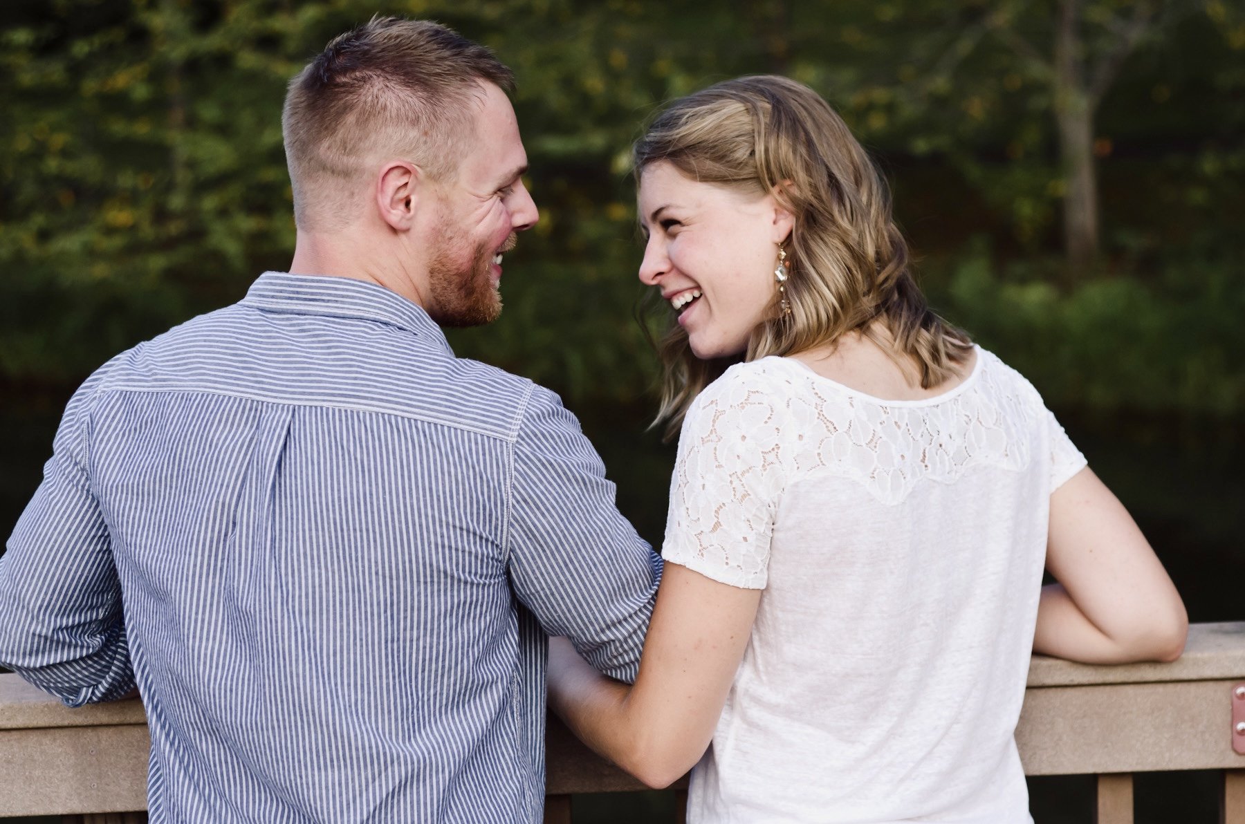 A young man and woman smiling at each other outdoors near a wooden railing with green trees in the background.
