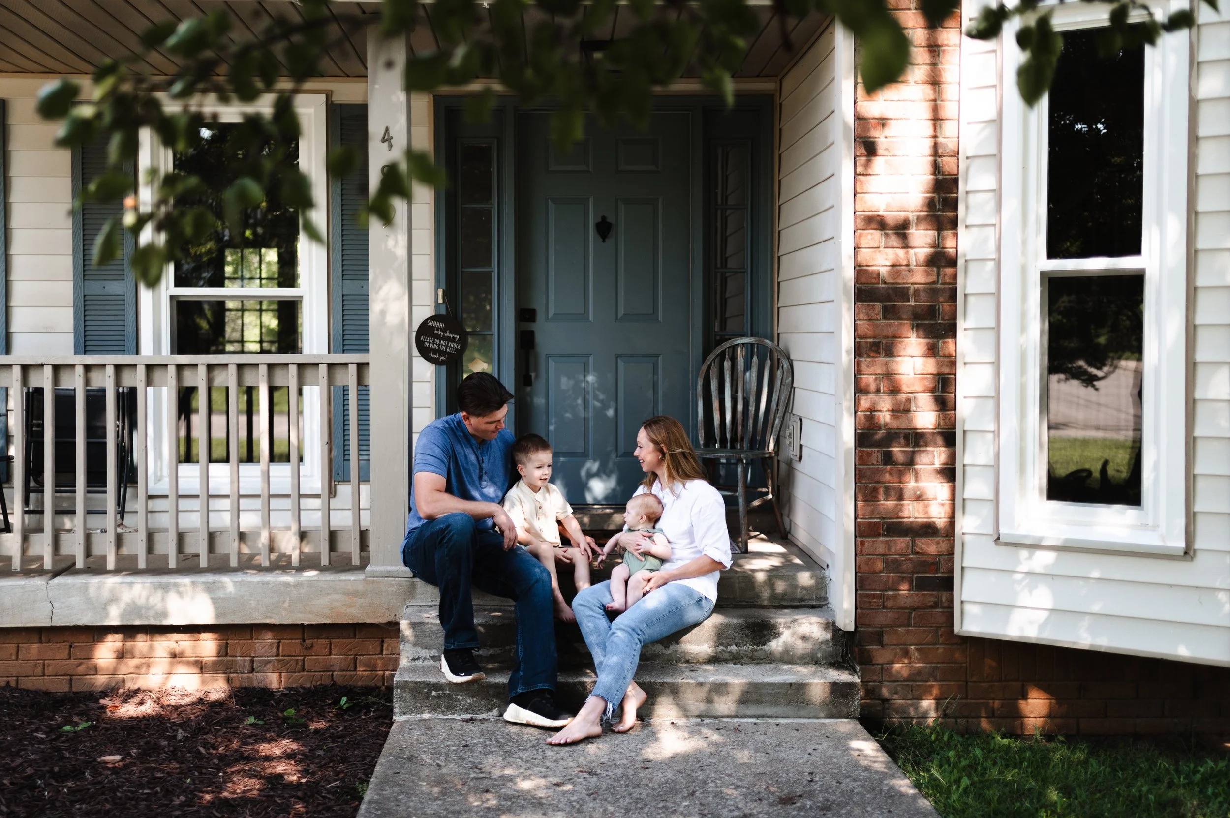 A family sitting on the front porch of a house, with two adults and three children, enjoying a conversation in the daytime.