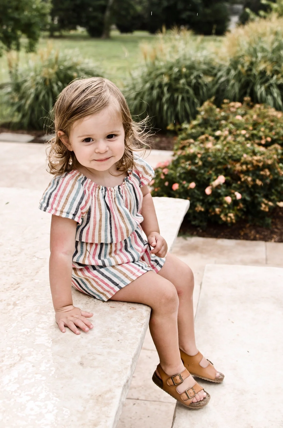 young brunette girl wearing striped romper and sandals sits on stone table in garden, clarksville tn photographer, nashville tn photographer, affordable, family photographer, milestone, birthday, outdoor, colorful, bright