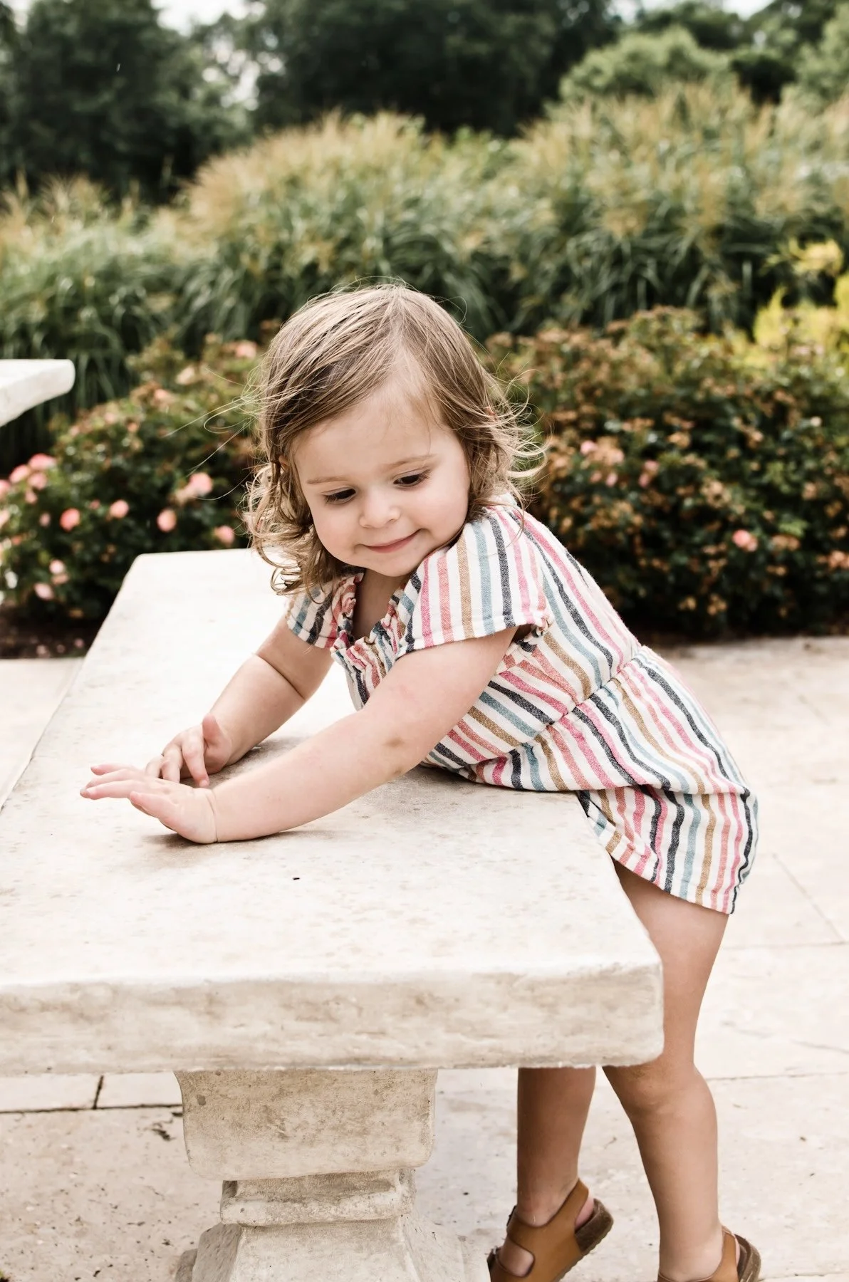 young girl wearing striped romper and sandals leans on stone bench in garden with flowers, clarksville tn photographer, nashville tn photographer, affordable, family photographer, milestone, birthday, outdoor, home session, garden portrait, spring