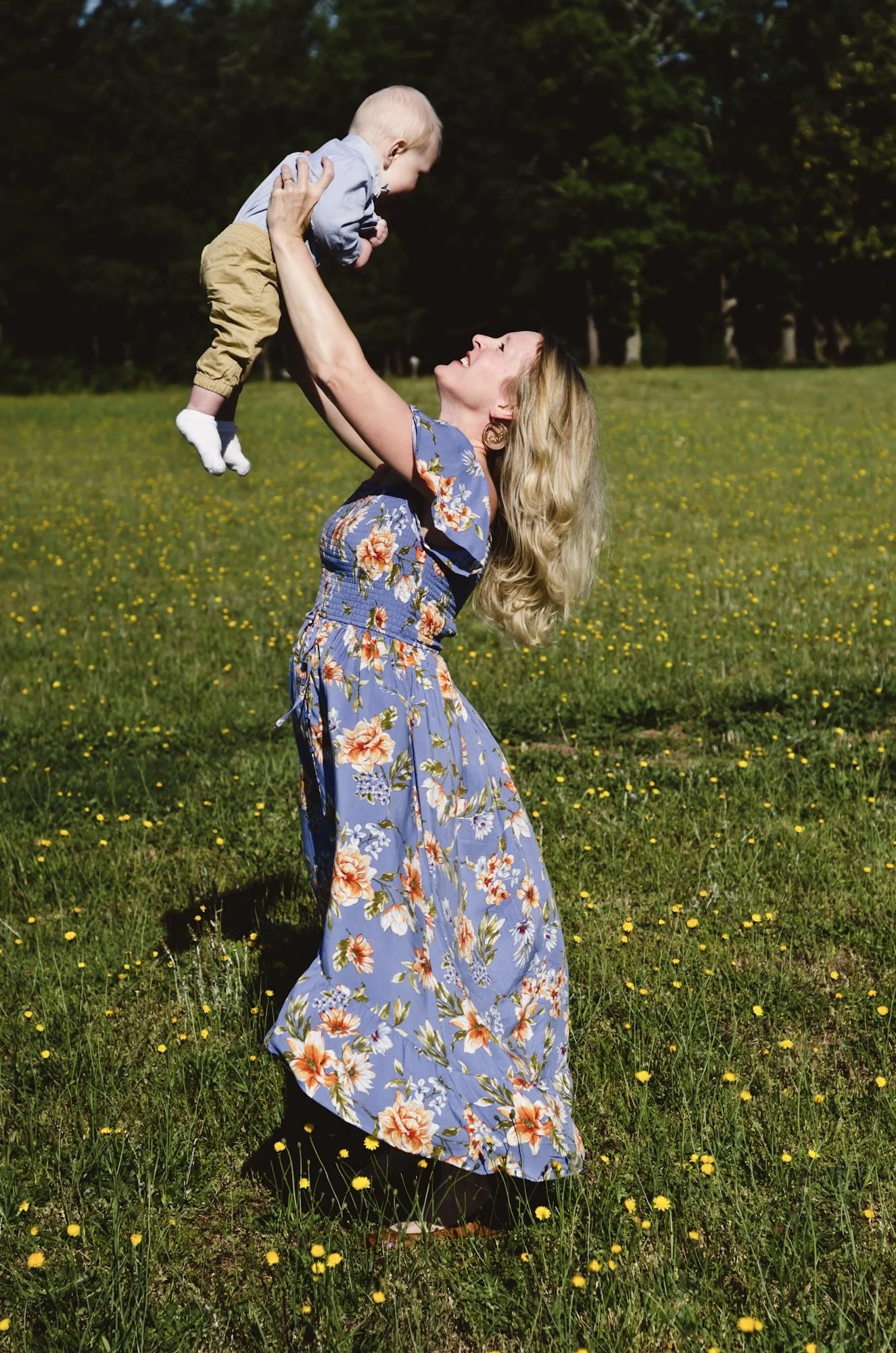blonde woman in blue and orange floral dress stands in yellow flower field on windy spring day while holding up baby boy, photographer, motherhood, milestone, affordable, clarksville tn, nashville tn, springfield tn, fort campbell ky