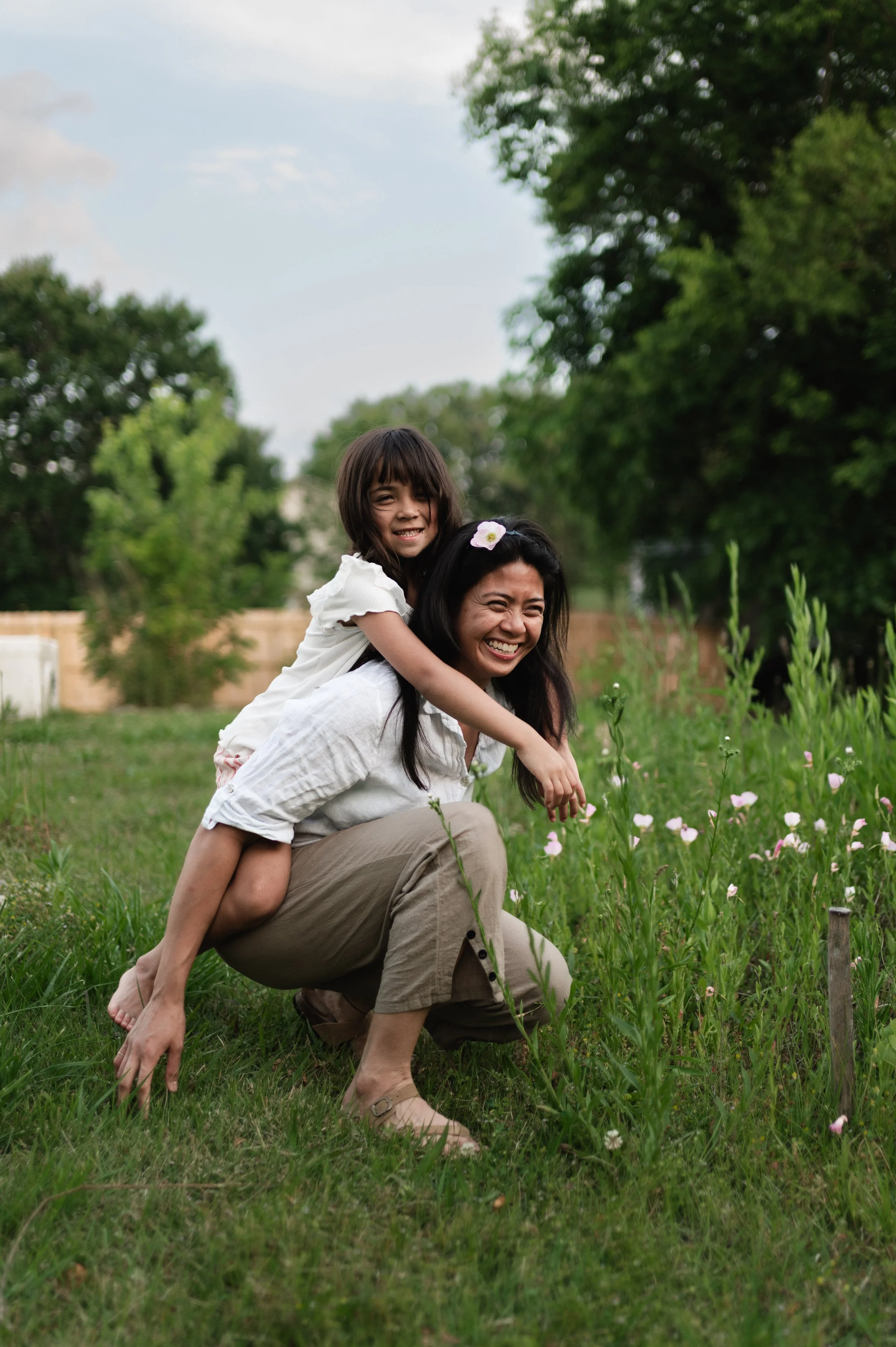 A woman and a young girl outdoors on a grassy field, smiling and playing. The woman is crouching while the girl is sitting on her back, with her arms around the woman's shoulders. There are trees and a fence in the background, and the girl has a flower in her hair.