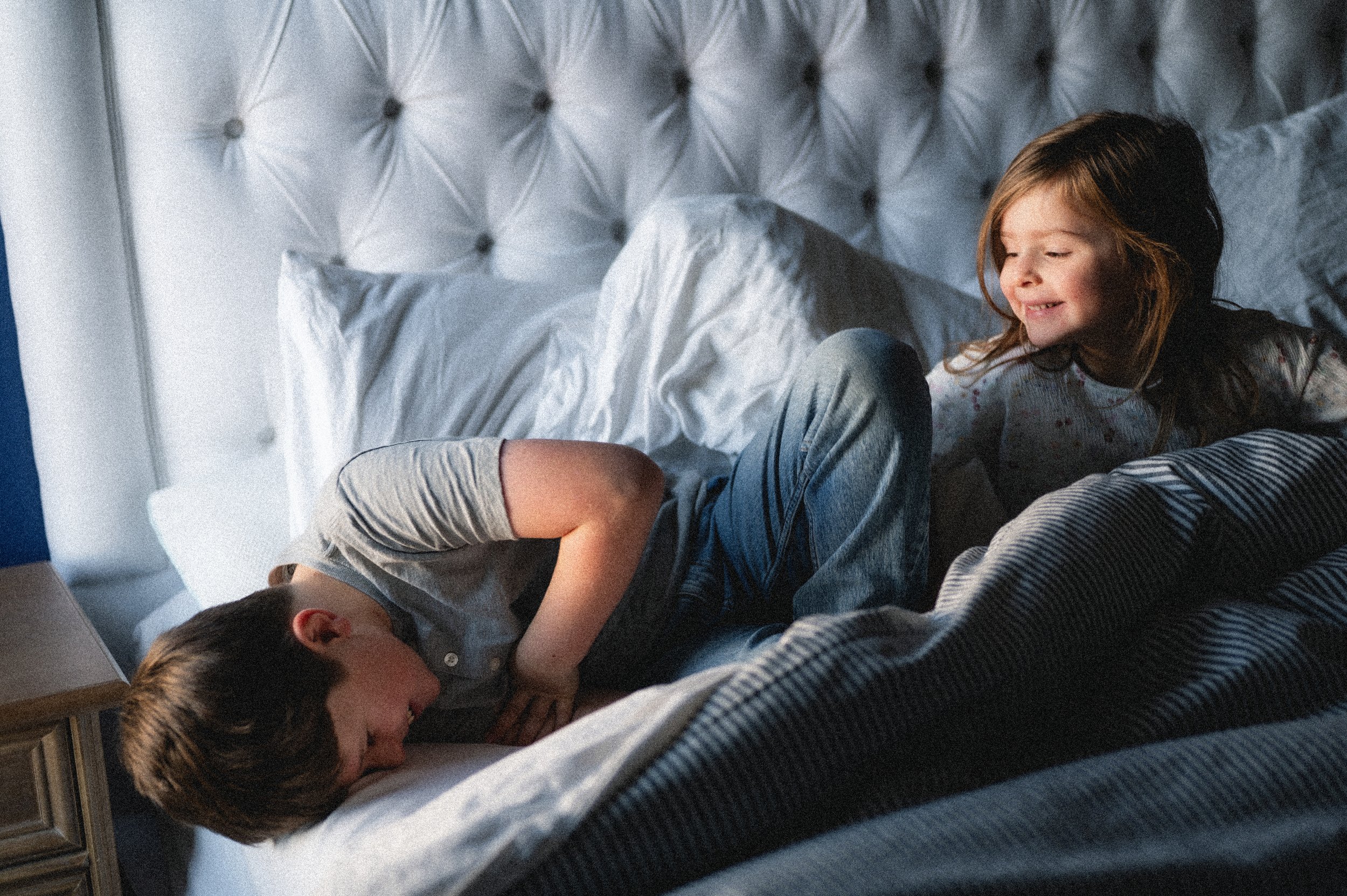 laughing and smiling young boy and girl sit in bed with white upholstered headboard, photographer, affordable, clarksville tn, nashville tn, documentary, lifestyle, moody, candid, in-home session, family photographer, brother and sister