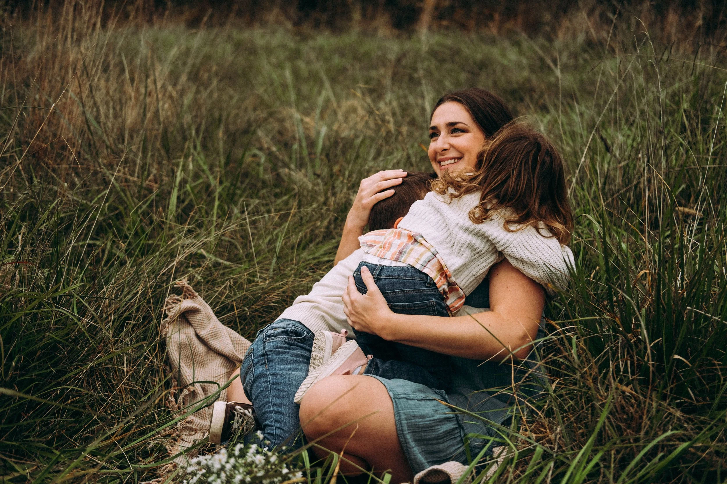A woman and two children hugging in a grassy field, with the woman smiling.
