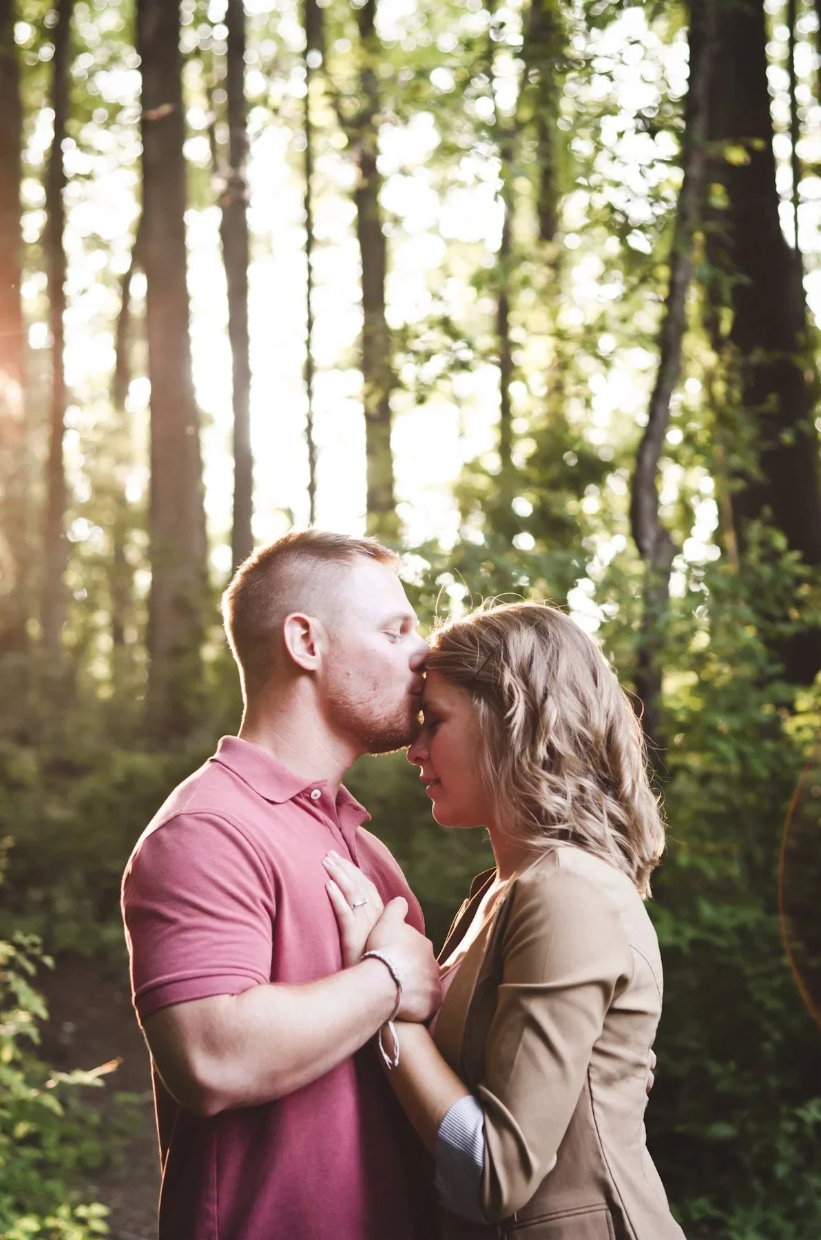 A man and woman in a forest, facing each other with eyes closed, the man gently kissing the woman's forehead, both holding hands and embracing.