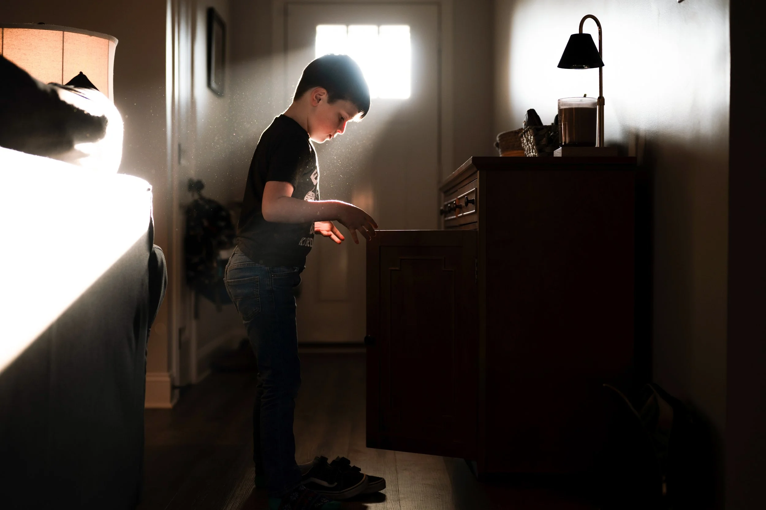 Dark haired boy stands in sun ray next to open cabinet in dark entryway of home, moody, clarksville tn, nashville tn, photographer, family photographer, in-home family session, dramatic, military family, documentary family photographer