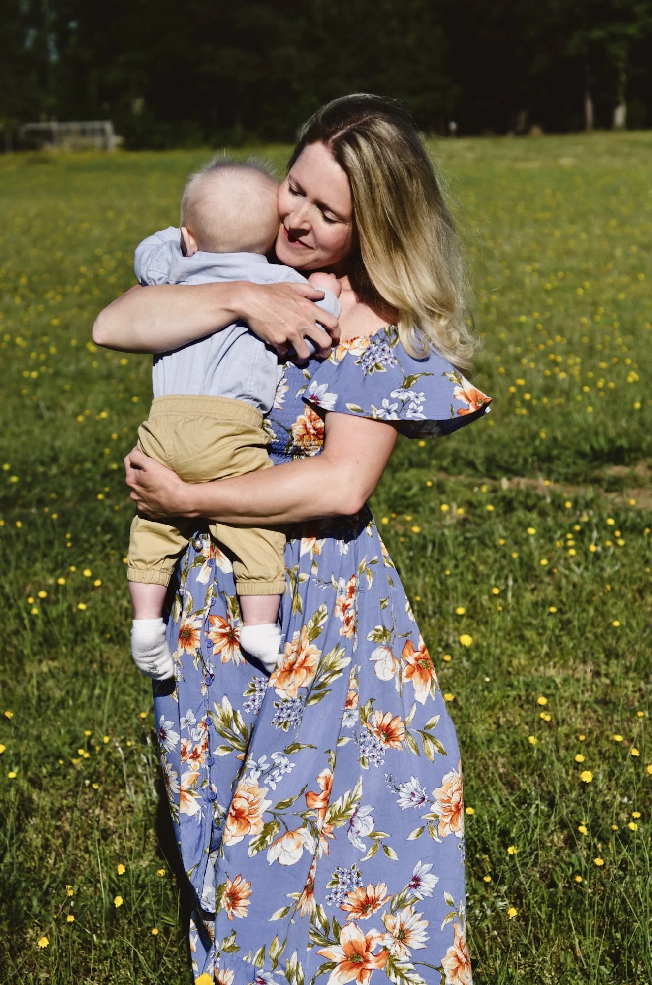 mother in floral blue dress holding baby walking through yellow flower field in bright sunlight on windy spring day, clarksville tn, nashville tn, photographer, motherhood, newborn photographer, family photographer, affordable
