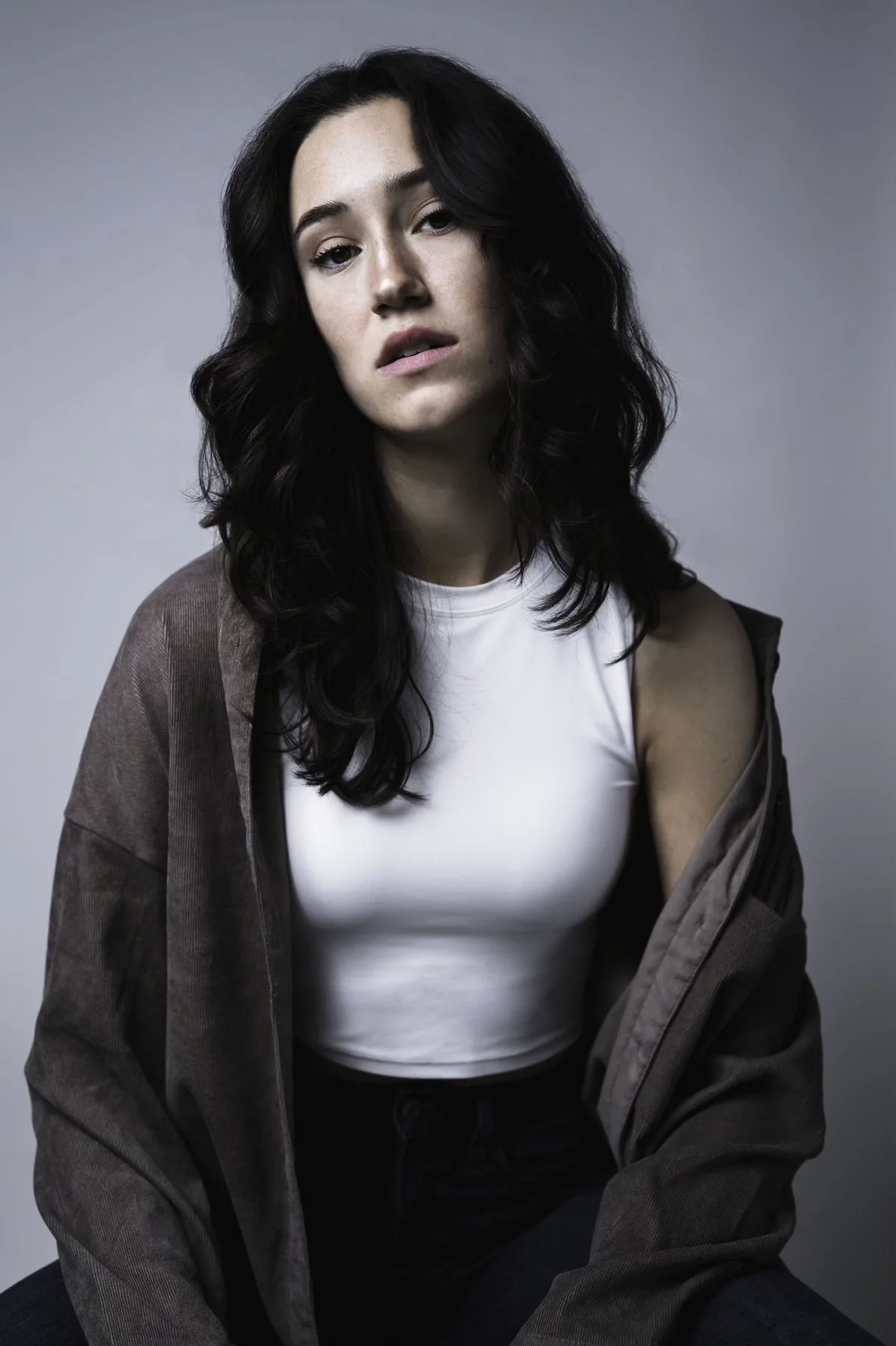 Portrait of a young woman with dark wavy hair wearing a white top and brown jacket against a gray background.