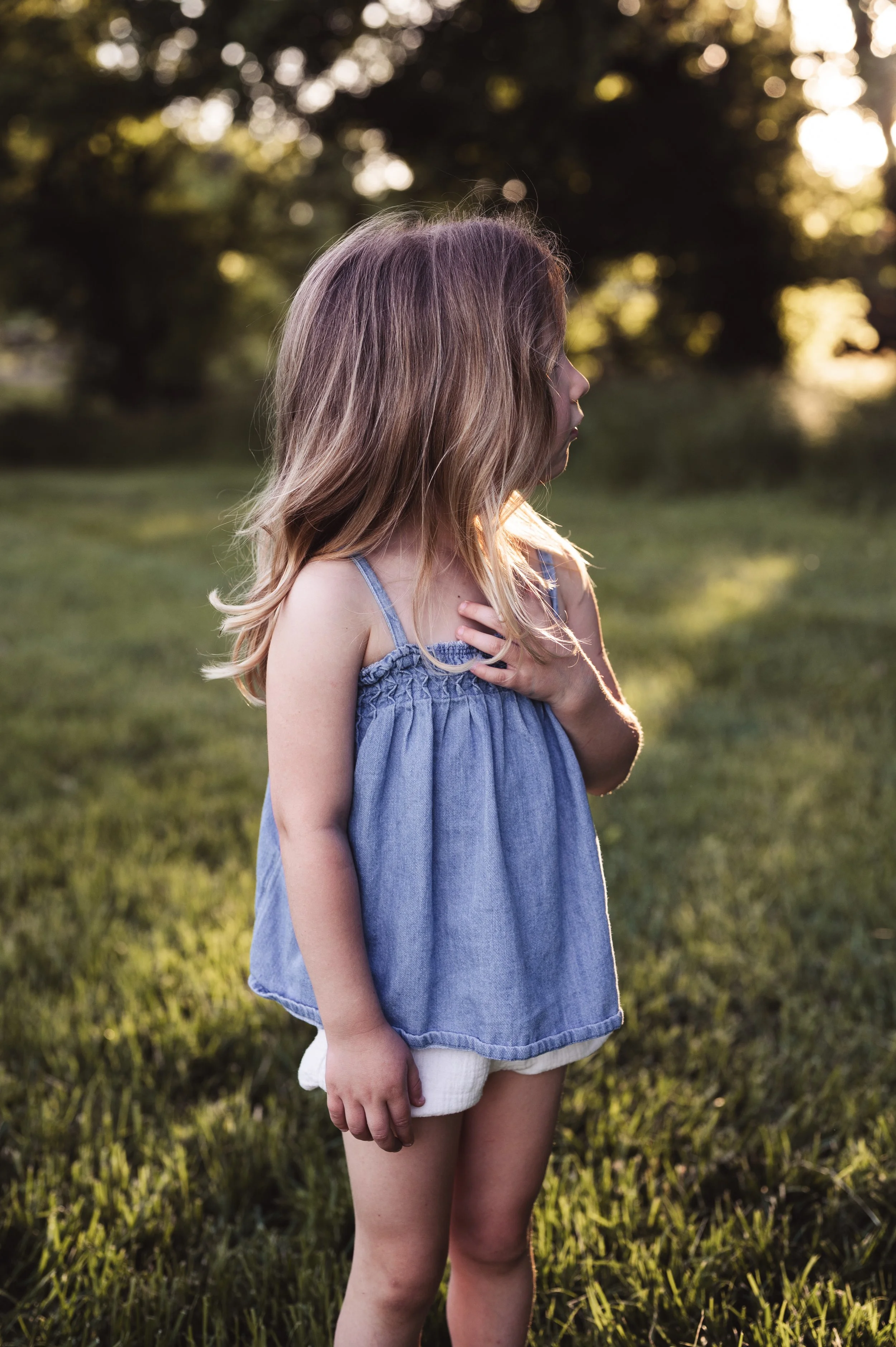 young blonde girl in blue shirt and white bloomers stands in grassy field with one had resting on chest looking toward trees, nashville tn, photographer, clarksville tn, family photographer, outdoor family photos, milestone