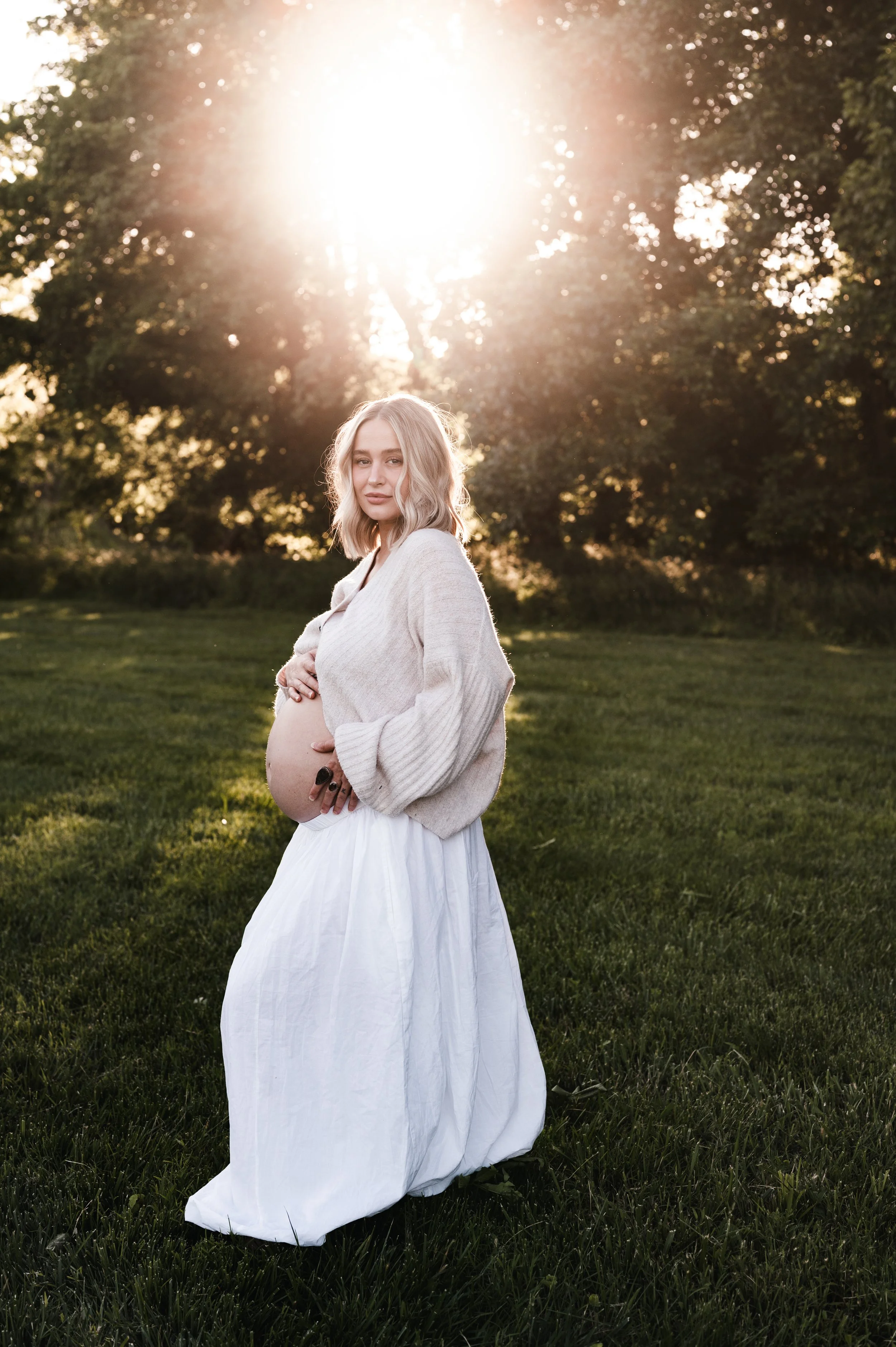 A pregnant woman in a white dress and beige sweater standing outdoors on a green lawn, with trees in the background and sunlight shining through the trees.