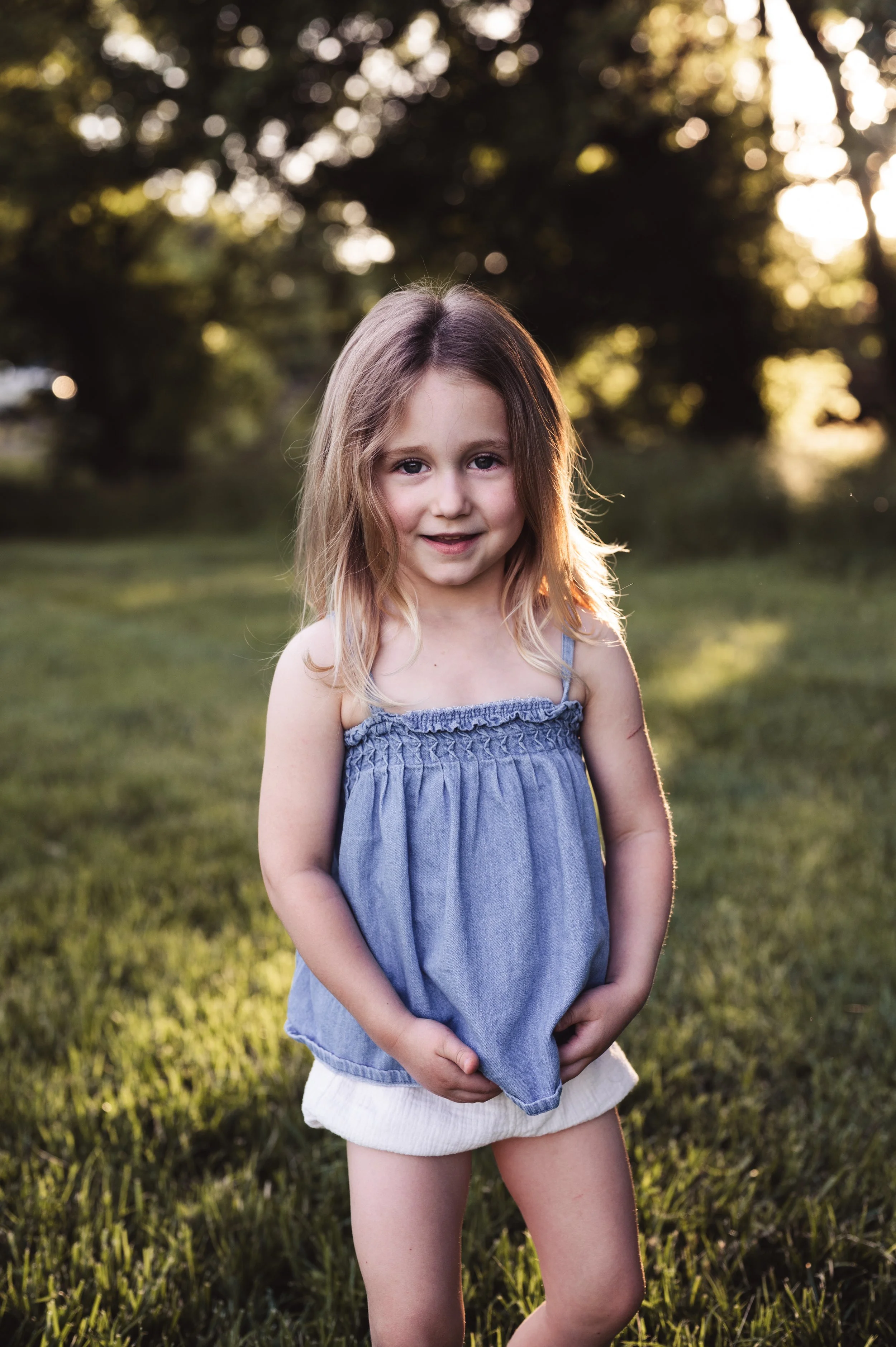 young blonde girl in blue blouse with white bloomers stands in grassy field with trees during summer sunset, clarksville tn, nashville tn, photographer, affordable, family photographer, milestone, outdoor portrait photographer
