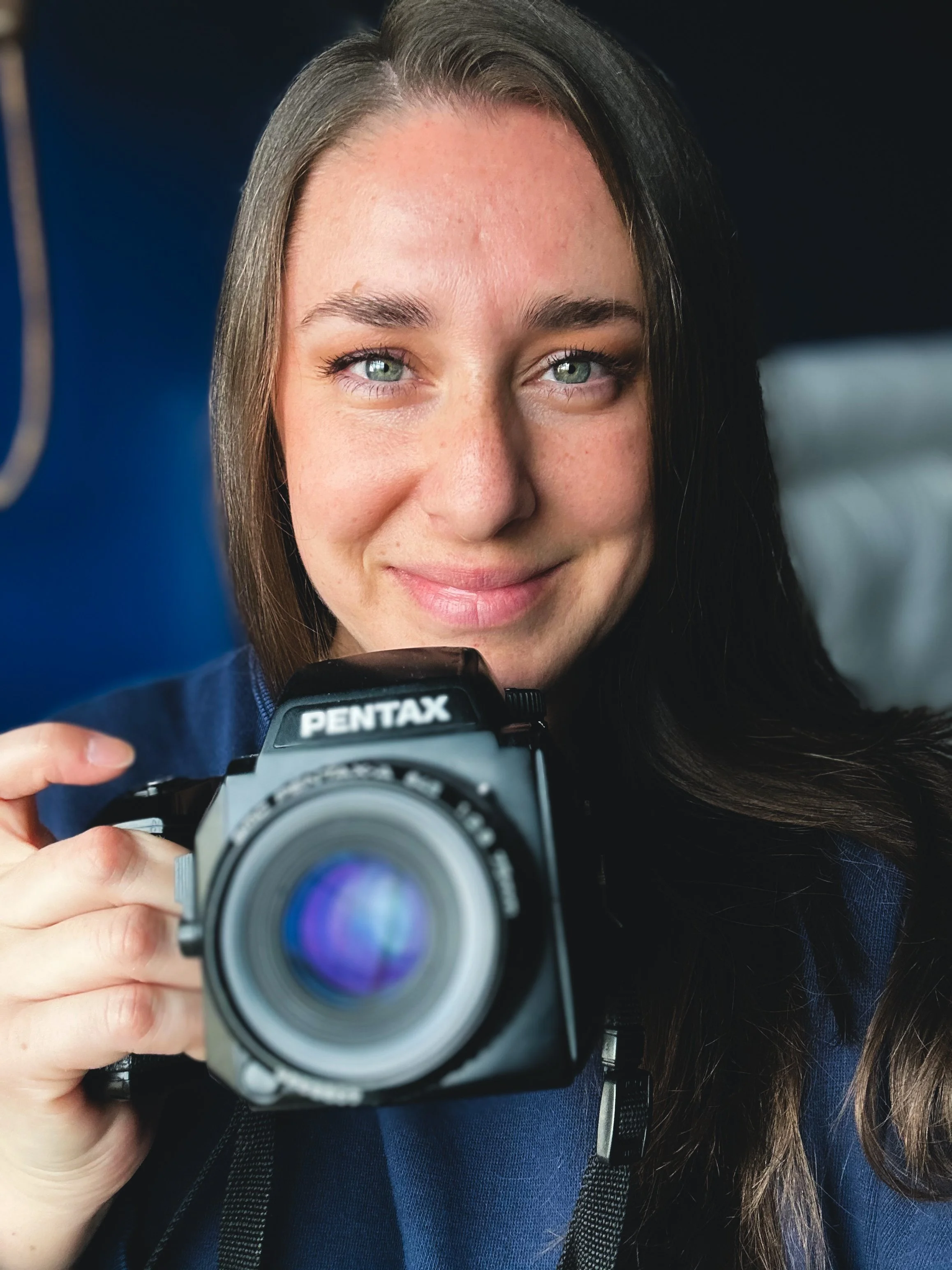 woman with bright blue eyes and long dark hair holds film camera in front of blurred navy background in Clarksville, Tennessee.