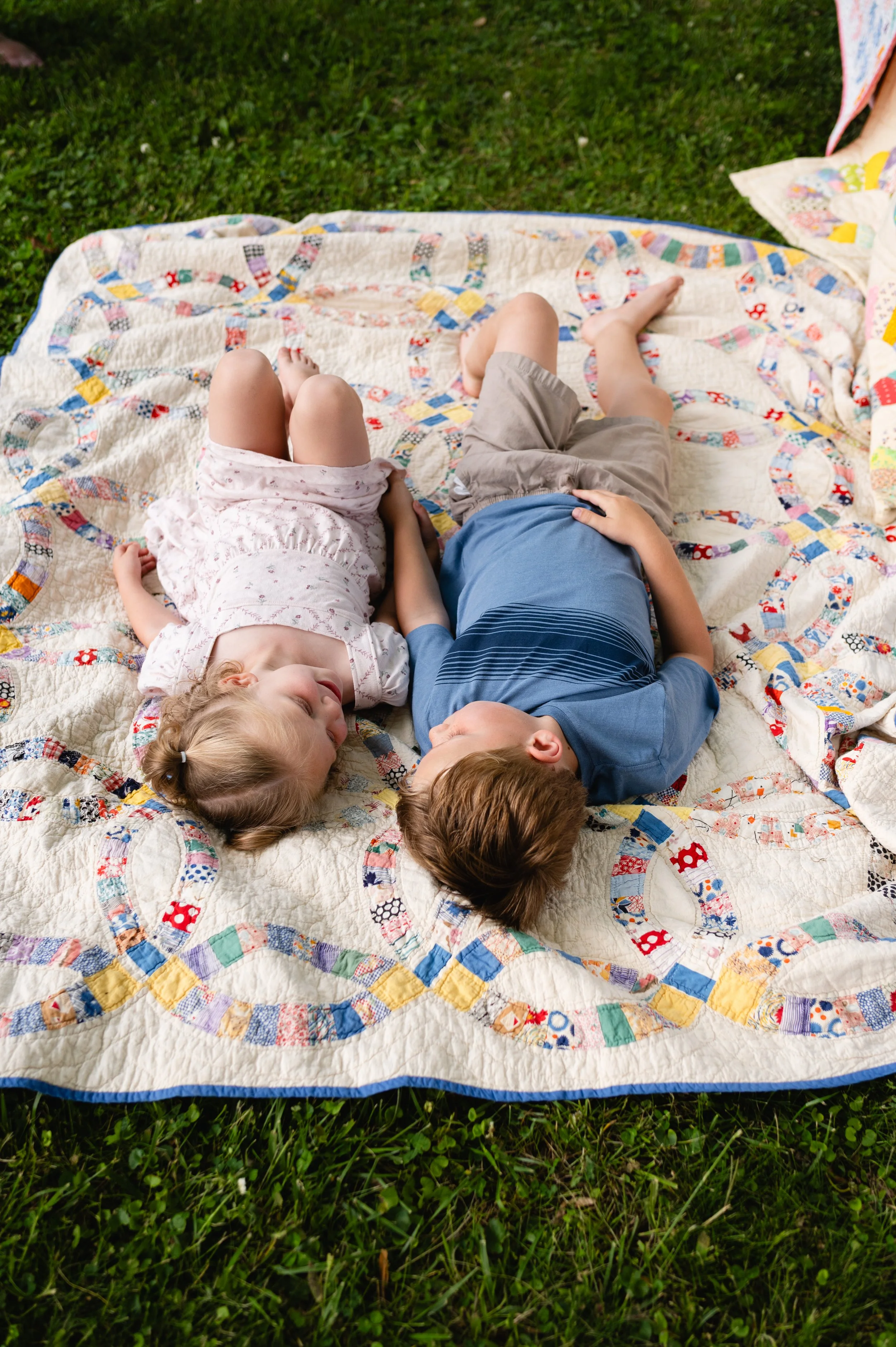 Two children lying on a colorful patchwork quilt on the grass, relaxing in an outdoor setting.