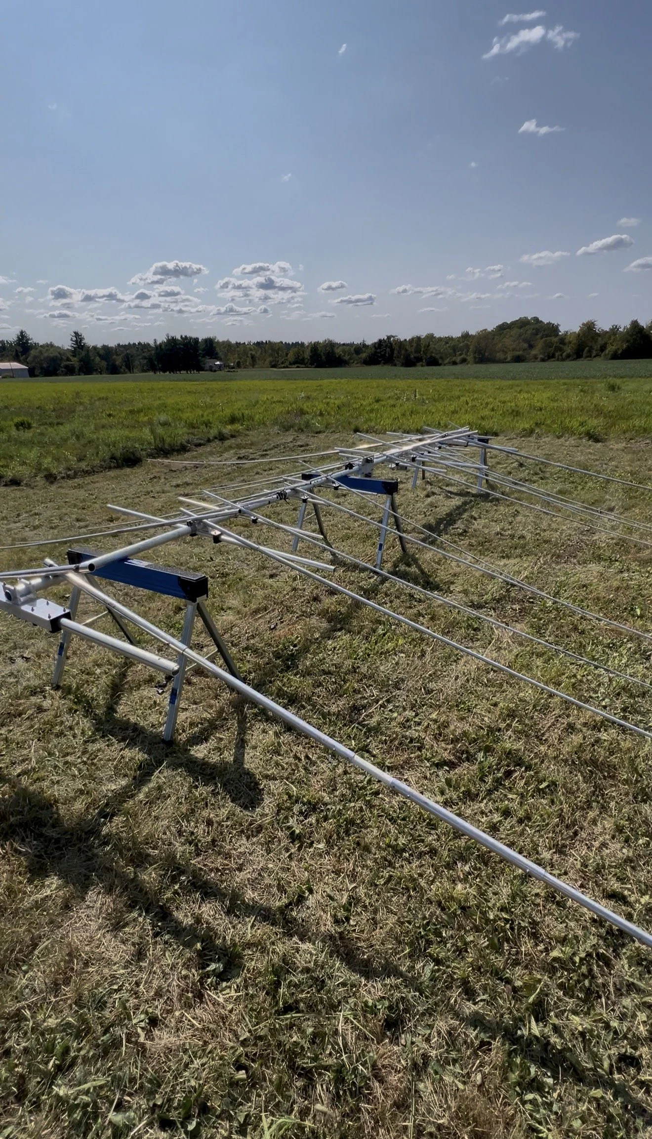 Sprinkler system set up in a grassy field with a clear blue sky and scattered clouds.